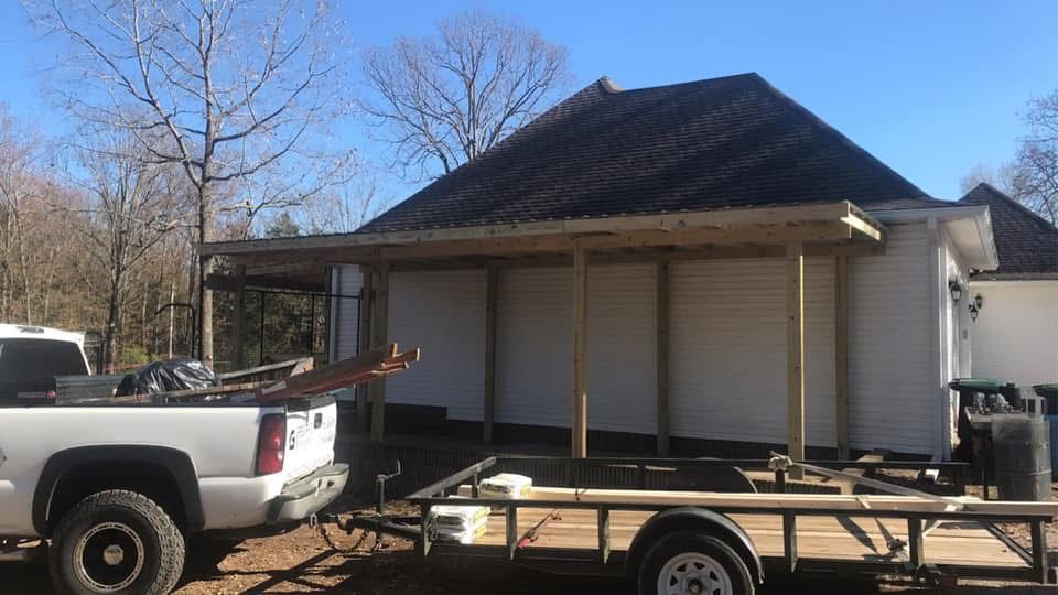 A pickup truck and trailer sit next to a house with an unfinished porch, on a sunny day.