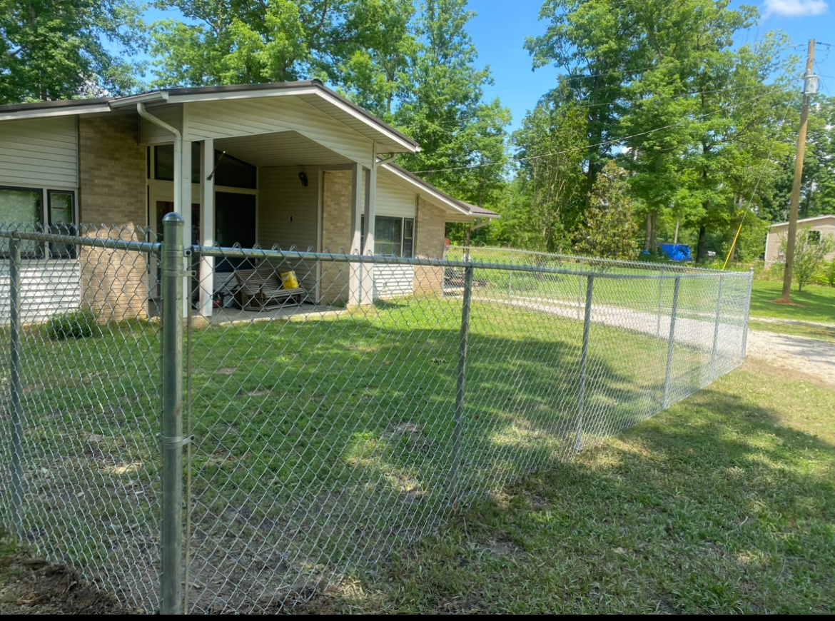 A house with a chain-link fence; green grass, trees, and blue sky.