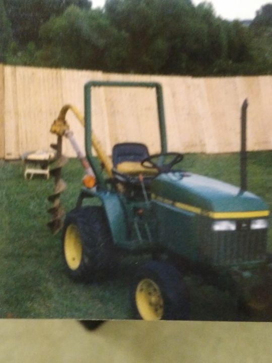 Green John Deere tractor with auger attached, on grass, next to a wooden fence.