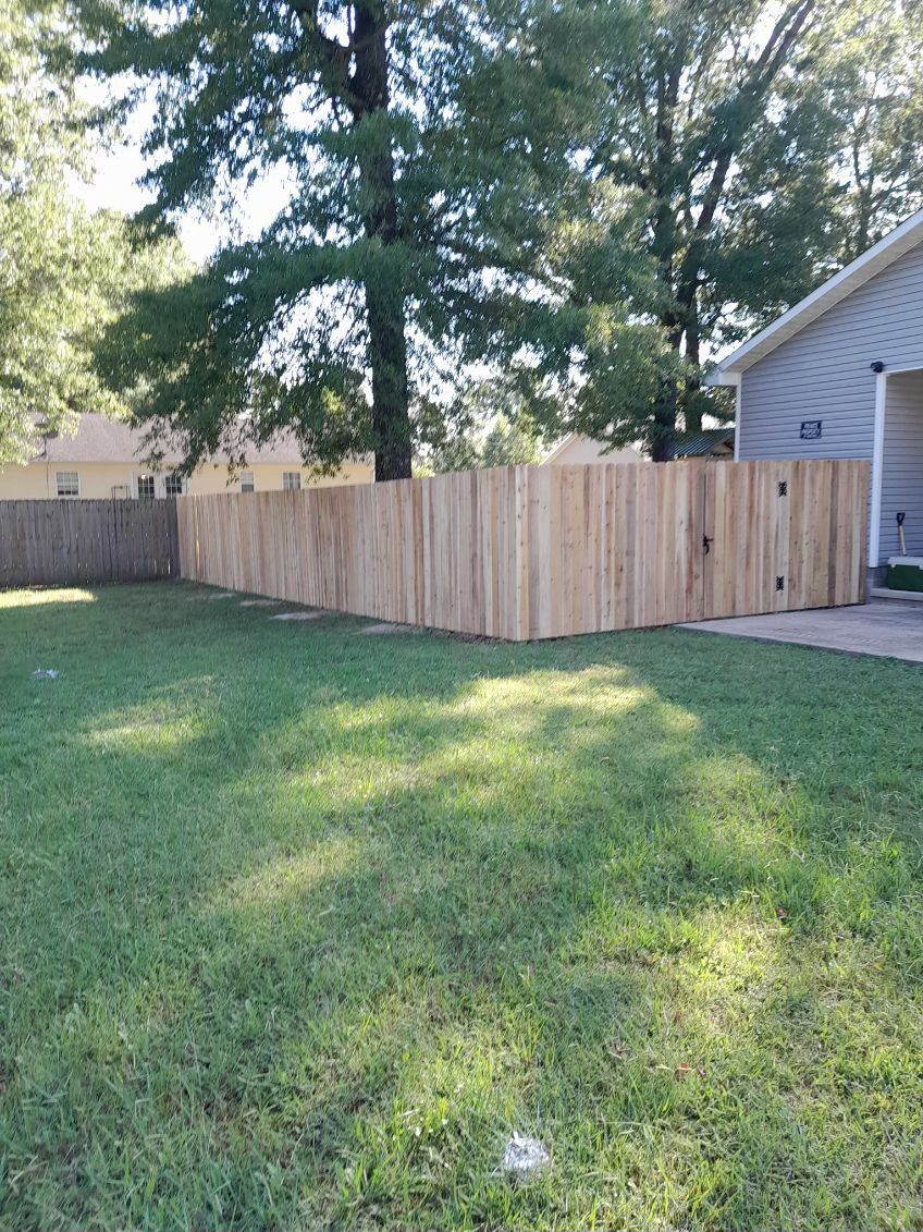 Wooden fence enclosing a grassy yard with a gate. Trees and a house are in the background.
