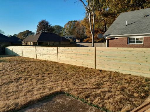 A newly constructed wooden fence surrounds a backyard with dry grass. Houses and trees are in the background.