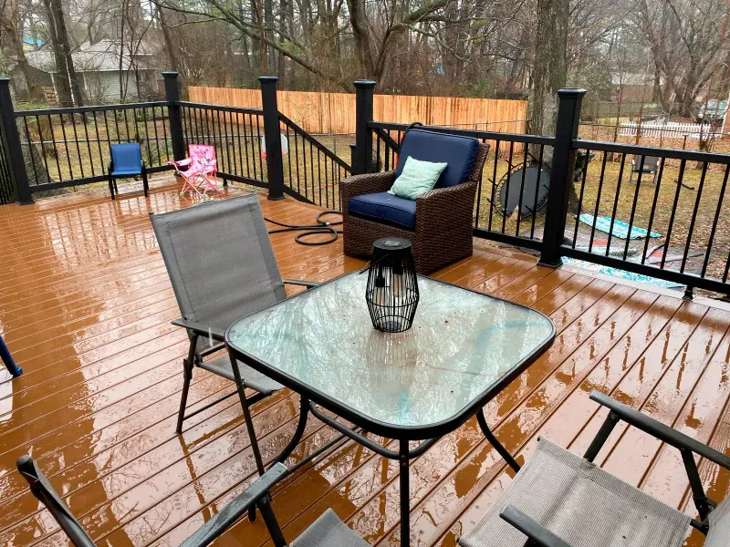 Outdoor deck with brown wooden planks, black railing, and patio furniture on a rainy day.