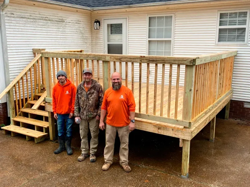 Three men stand in front of a new wooden deck attached to a white house.