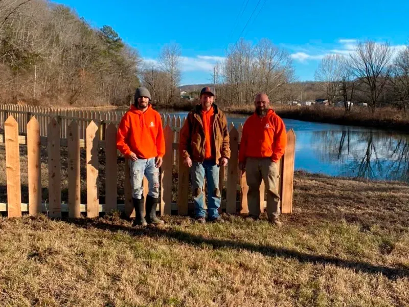 Three men in orange shirts and jeans stand near a wooden fence by a pond on a sunny day.