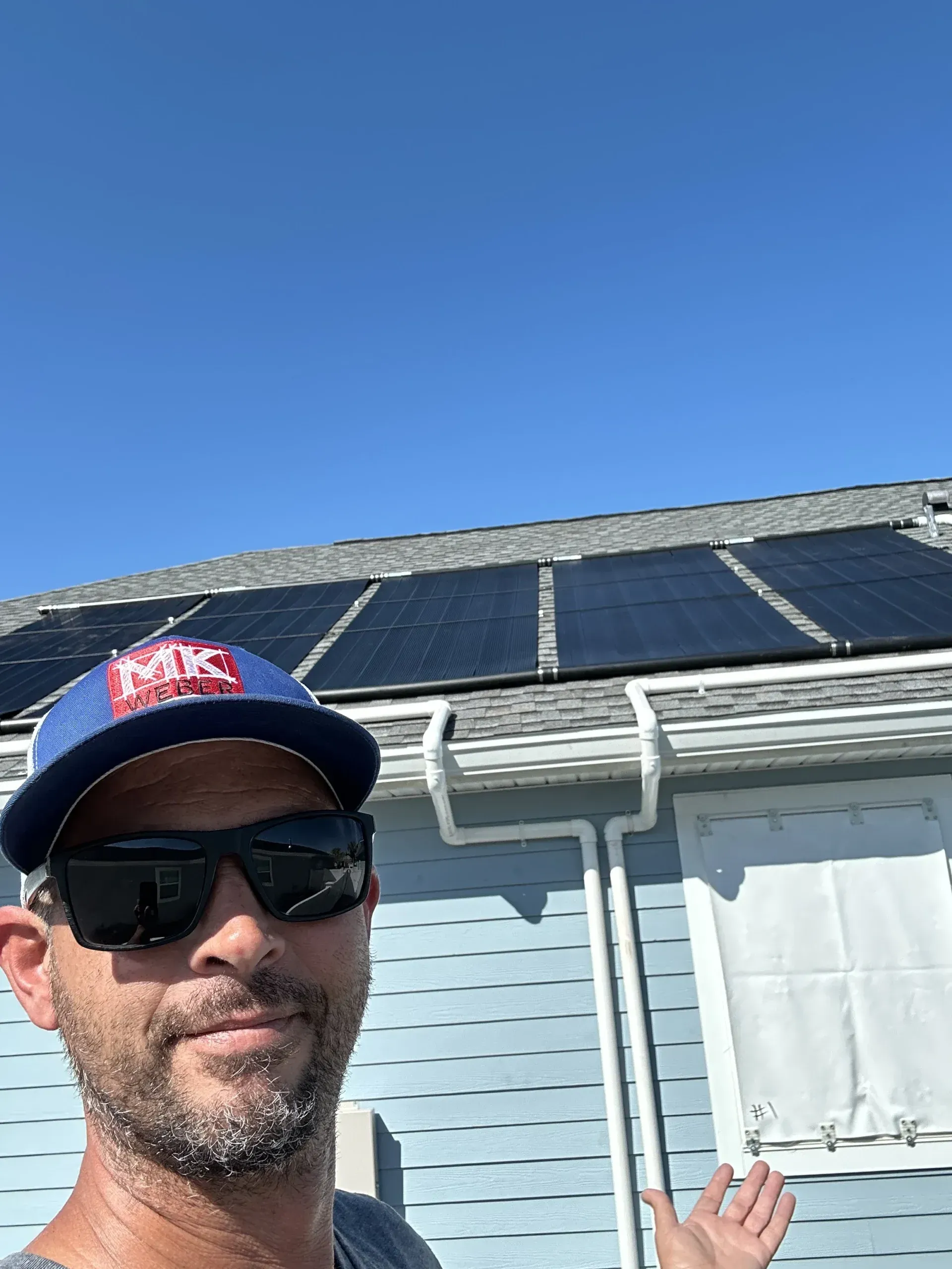 Man in sunglasses and cap, points to a roof with solar panels, blue siding and sky.