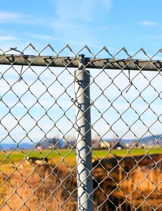 Chain-link fence with a metal post, in front of a field and blue sky.