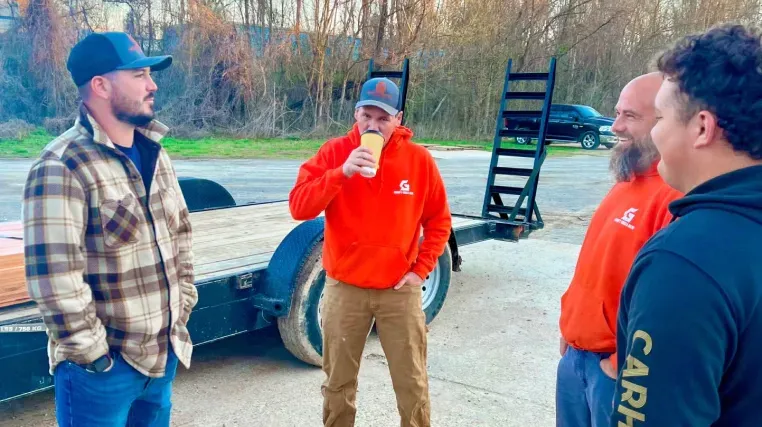 Four men in casual attire near a trailer. One drinks from a cup. Outdoors.