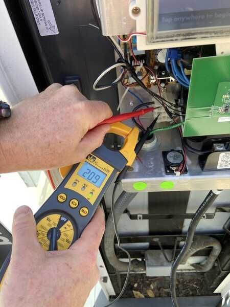 Hands using a clamp meter to test wires inside a machine. The meter reads 209.
