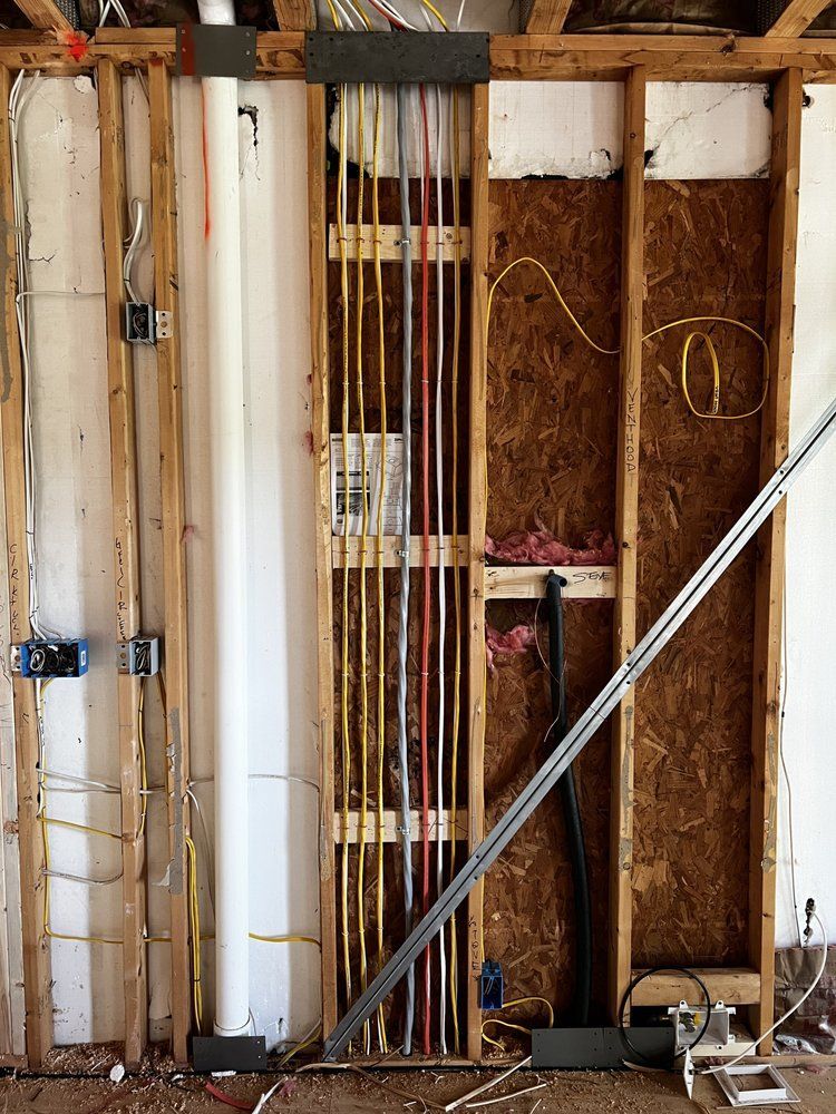 Interior wall under construction, showing electrical wiring in various colors, exposed studs, and a white pipe.