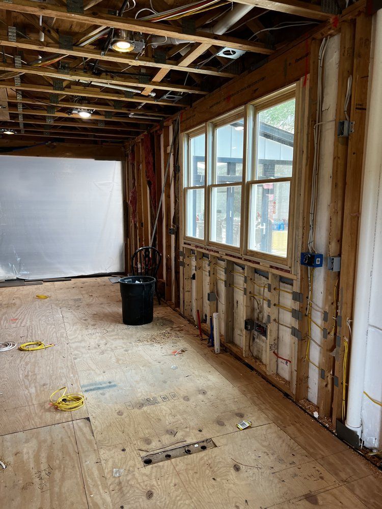 Interior of a room under renovation with exposed beams, wooden frames, windows, wiring, and debris.