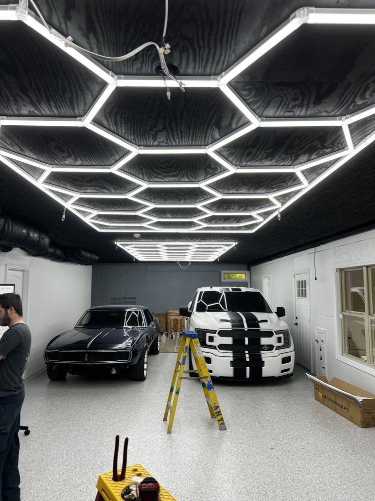 A man in a garage with two cars under hexagon-shaped lights. White truck with black stripes, black car.