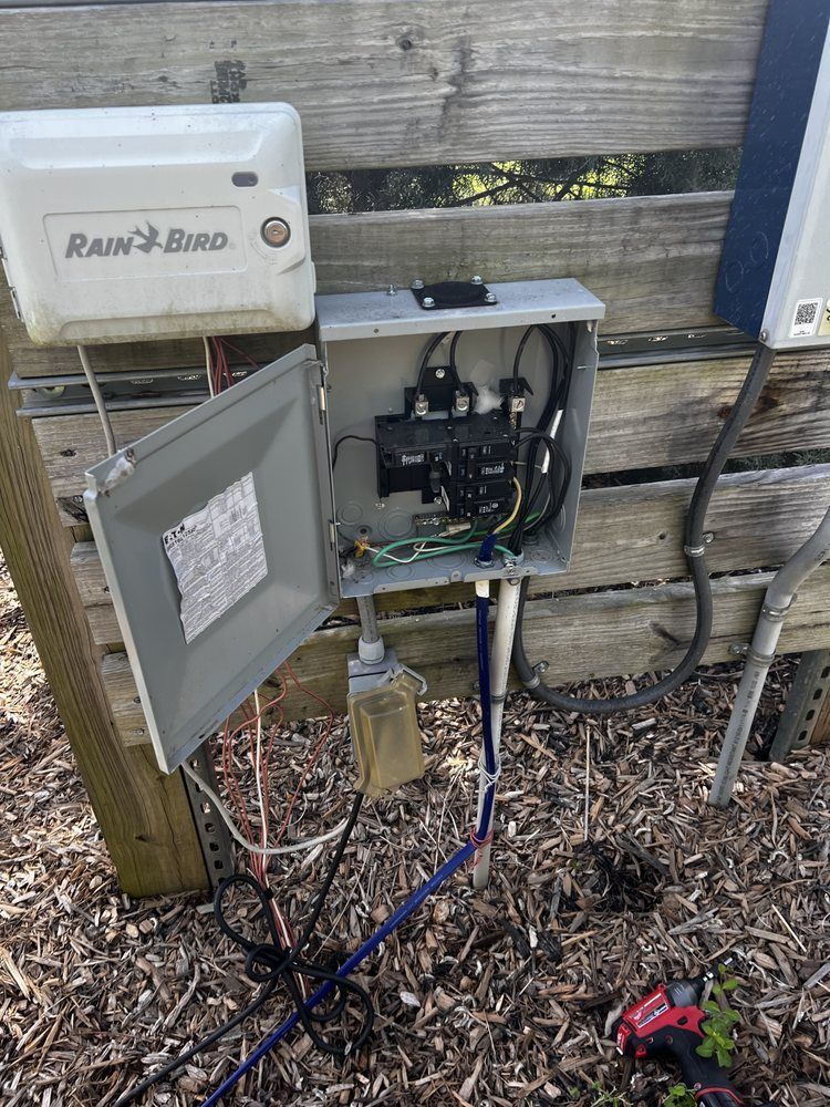 Open electrical box with wires, a Rain Bird controller, and surrounding wood and ground.