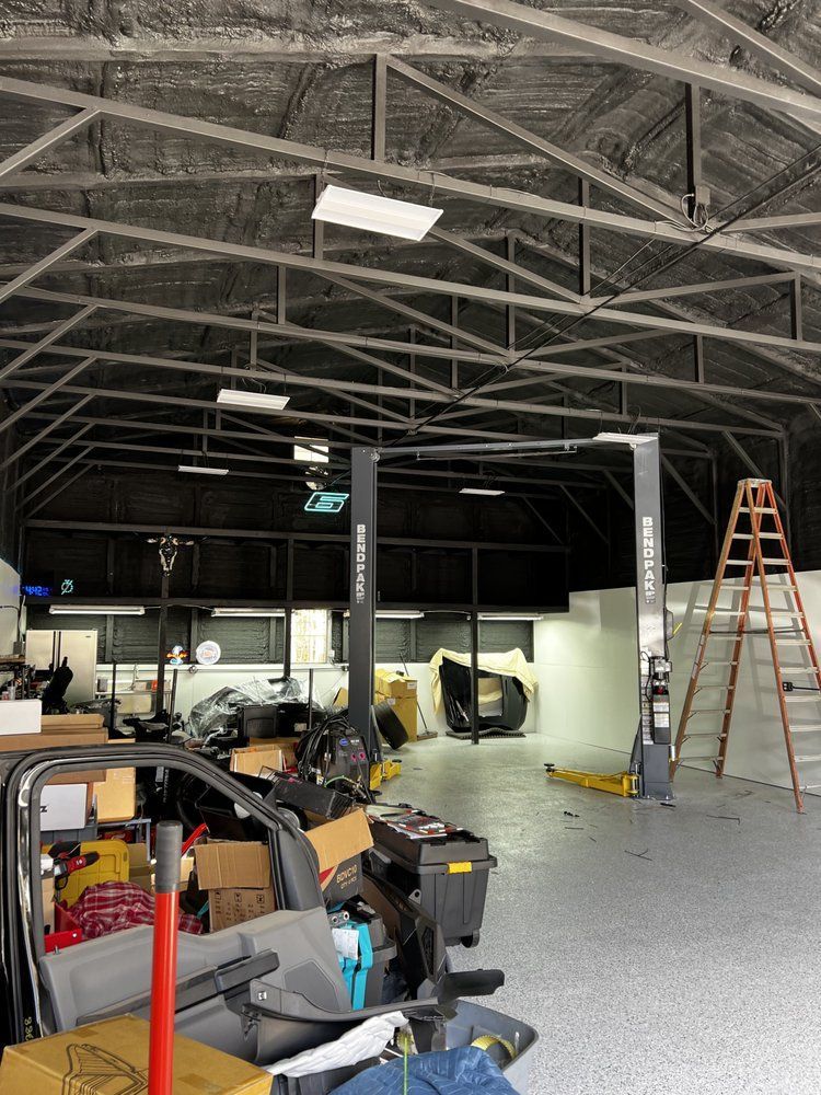 A cluttered garage interior with a car lift, ladder, and a partially visible vehicle. Gray, black, and white tones dominate the space.