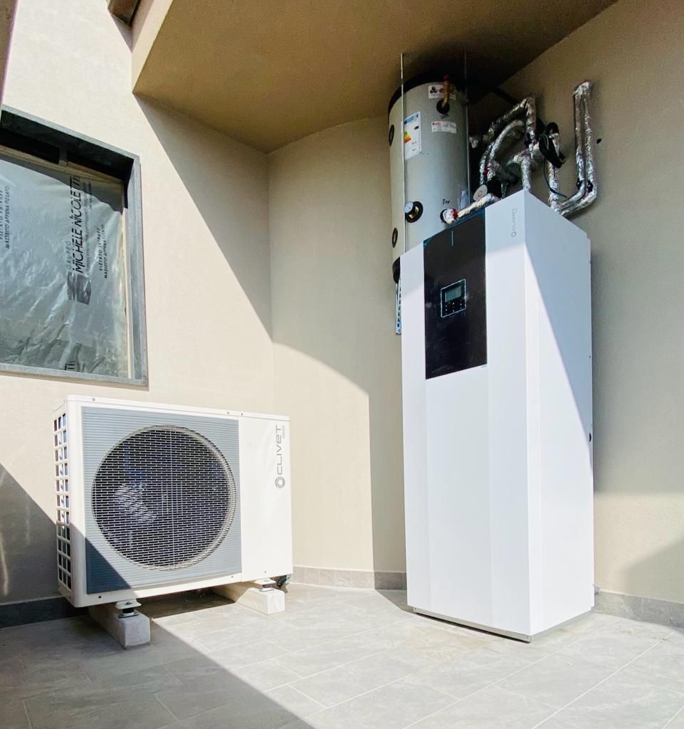 Un sistema di pompa di calore esterna con condensatore, scaldabagno e unità interna contro una parete beige.