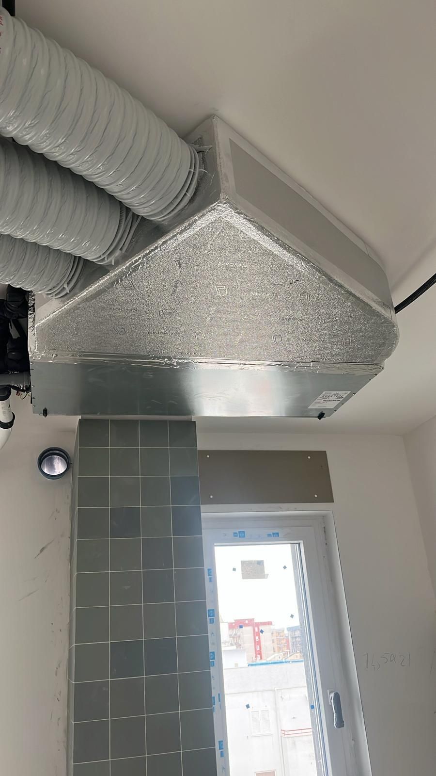 Cappa da cucina con più condotti di scarico argentati. La cappa è posizionata sopra un paraschizzi piastrellato e una finestra.