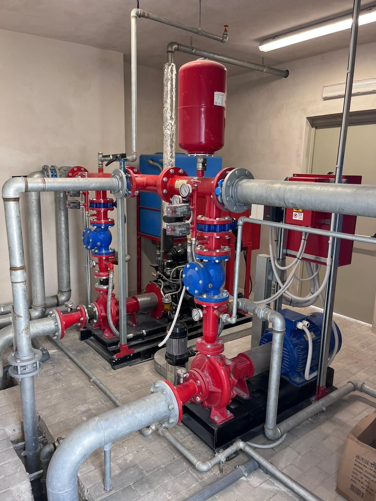 Impianto idraulico con valvole rosse e blu, tubi, pompe e un serbatoio rosso in una stanza.