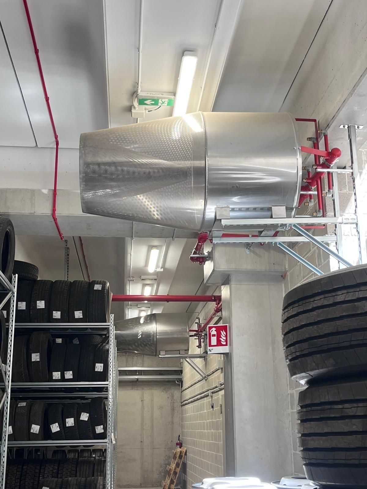 Grande serbatoio metallico, probabilmente per la soppressione degli incendi, montato su un'impalcatura in un magazzino, con pneumatici in primo piano.