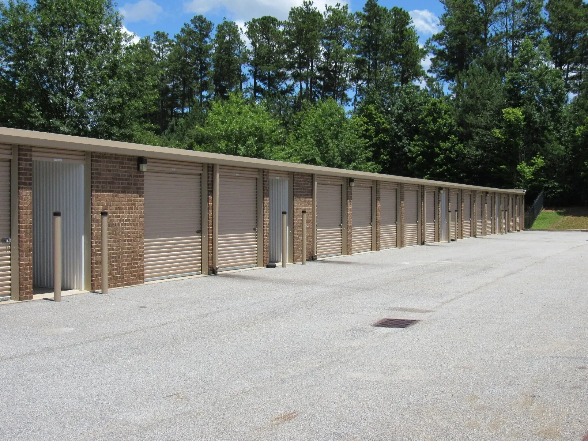Storage units with beige doors and brown brick trim, set on a gravel lot with trees in the background.