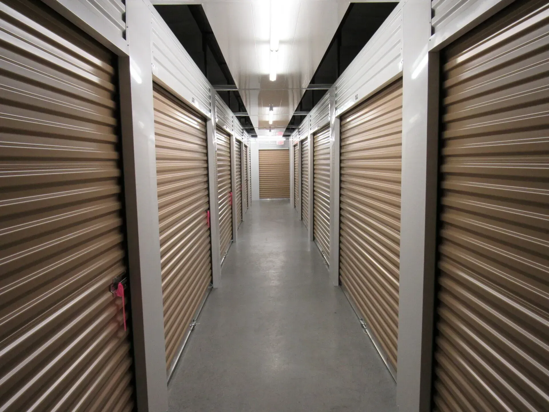 Indoor hallway of storage units; tan roller doors, white frames, fluorescent lights.