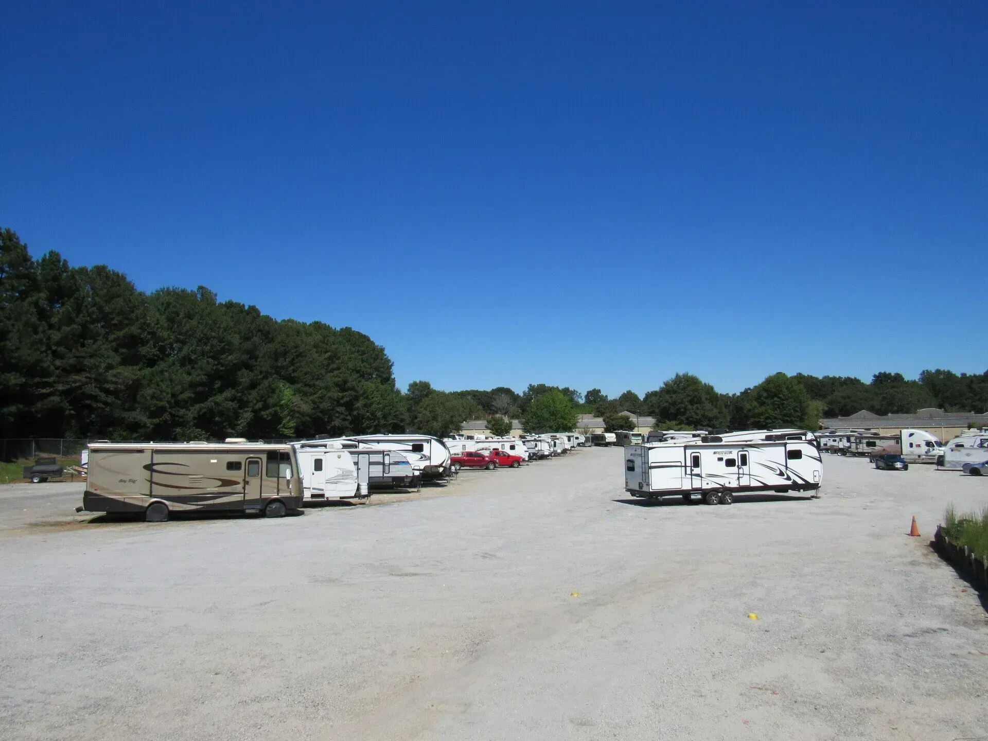RV storage lot under a clear blue sky; several RVs parked on gravel. Trees in background.