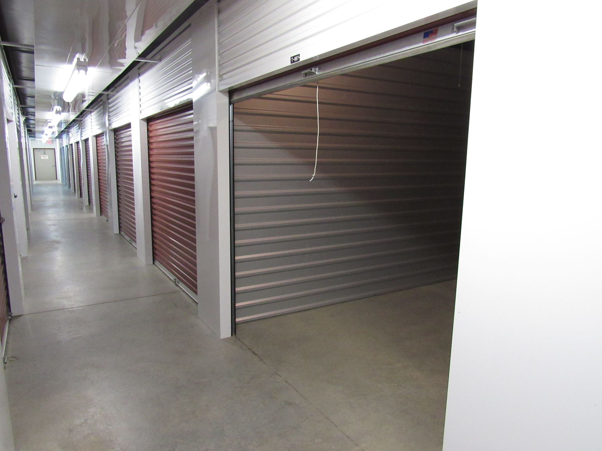 Storage units with tan doors line a black asphalt drive under a clear blue sky.