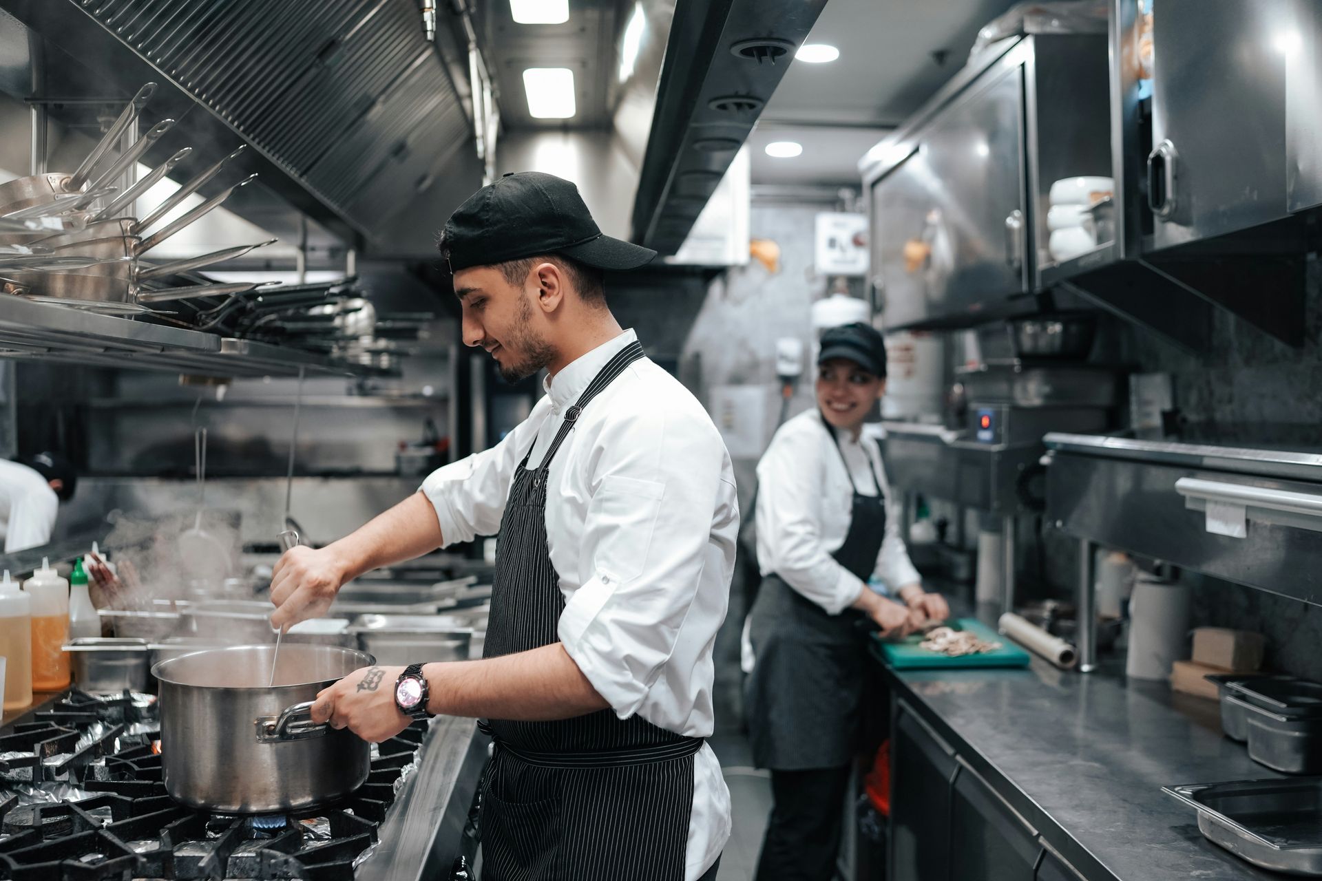 Two chefs working in a commercial kitchen; one stirring a pot on the stove, the other chopping vegetables.