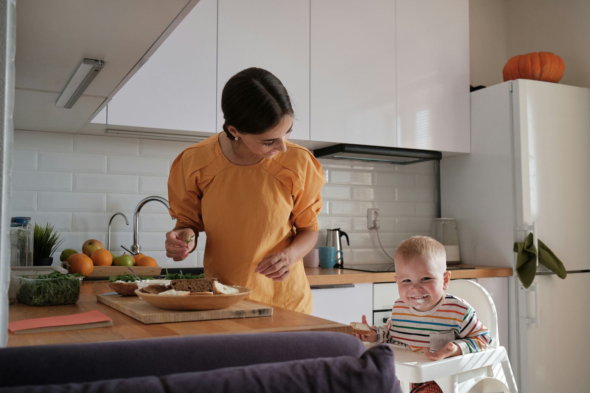 Woman in orange top prepares food with a toddler in a high chair in a kitchen.