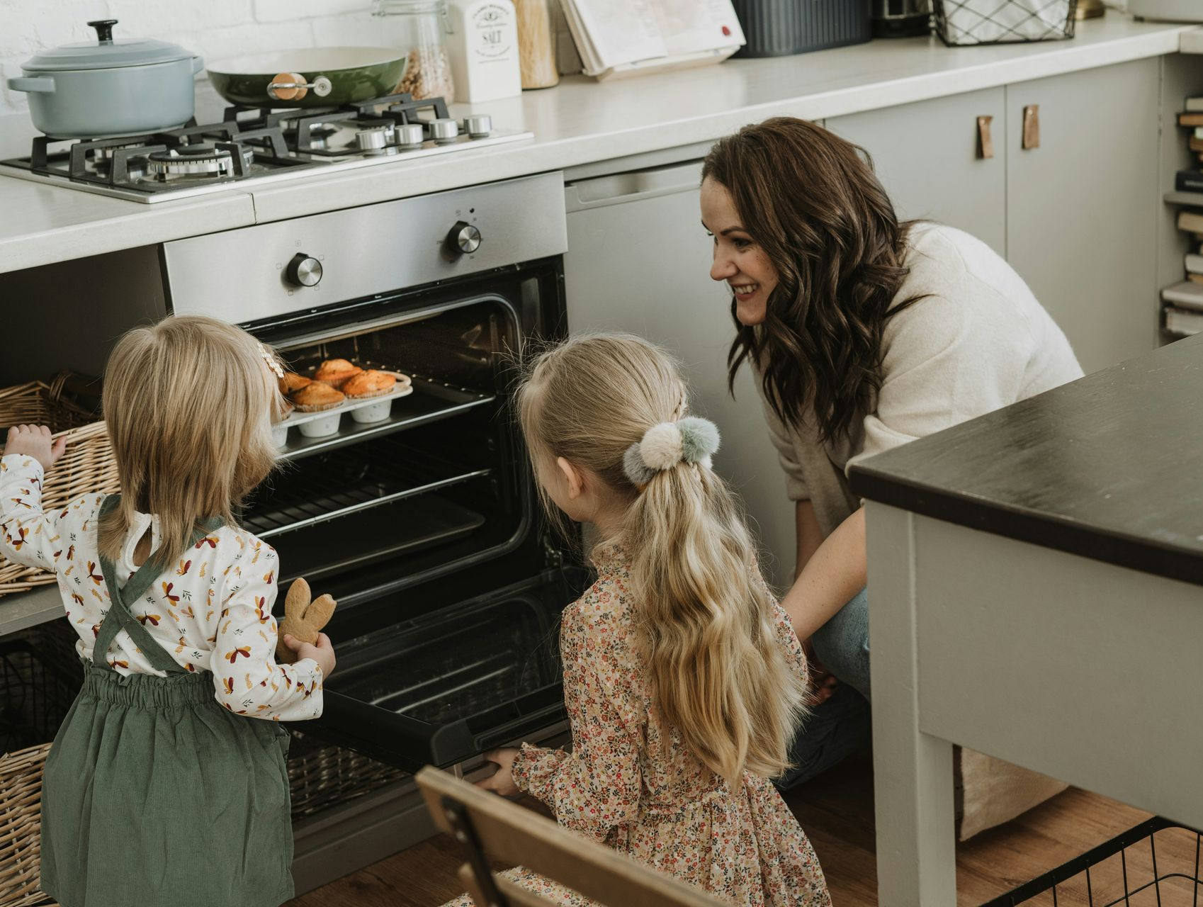 Woman smiles as she helps two young children take muffins from oven in a kitchen.