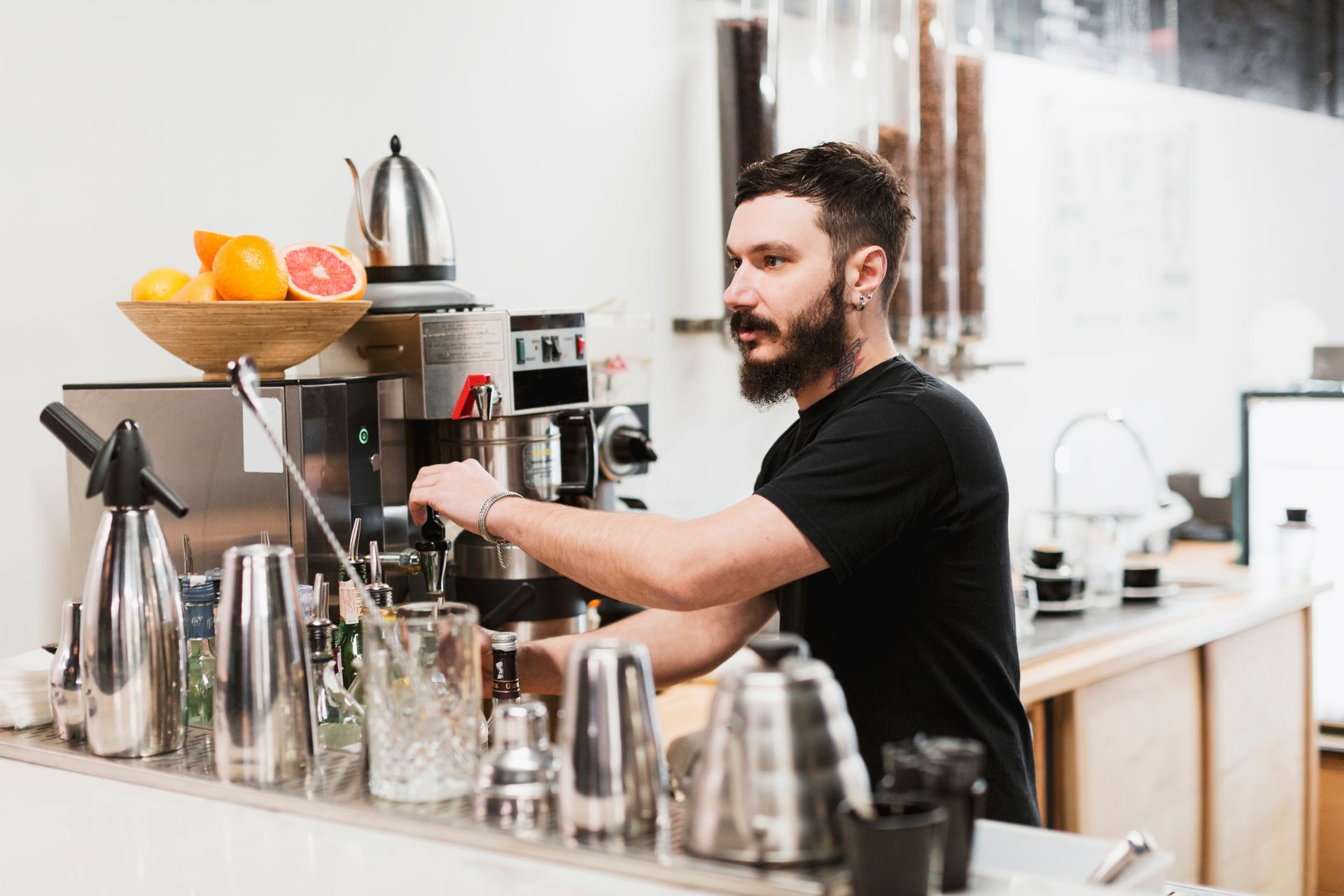 A barista with a beard operates an espresso machine behind a counter. Citrus fruit in a bowl.