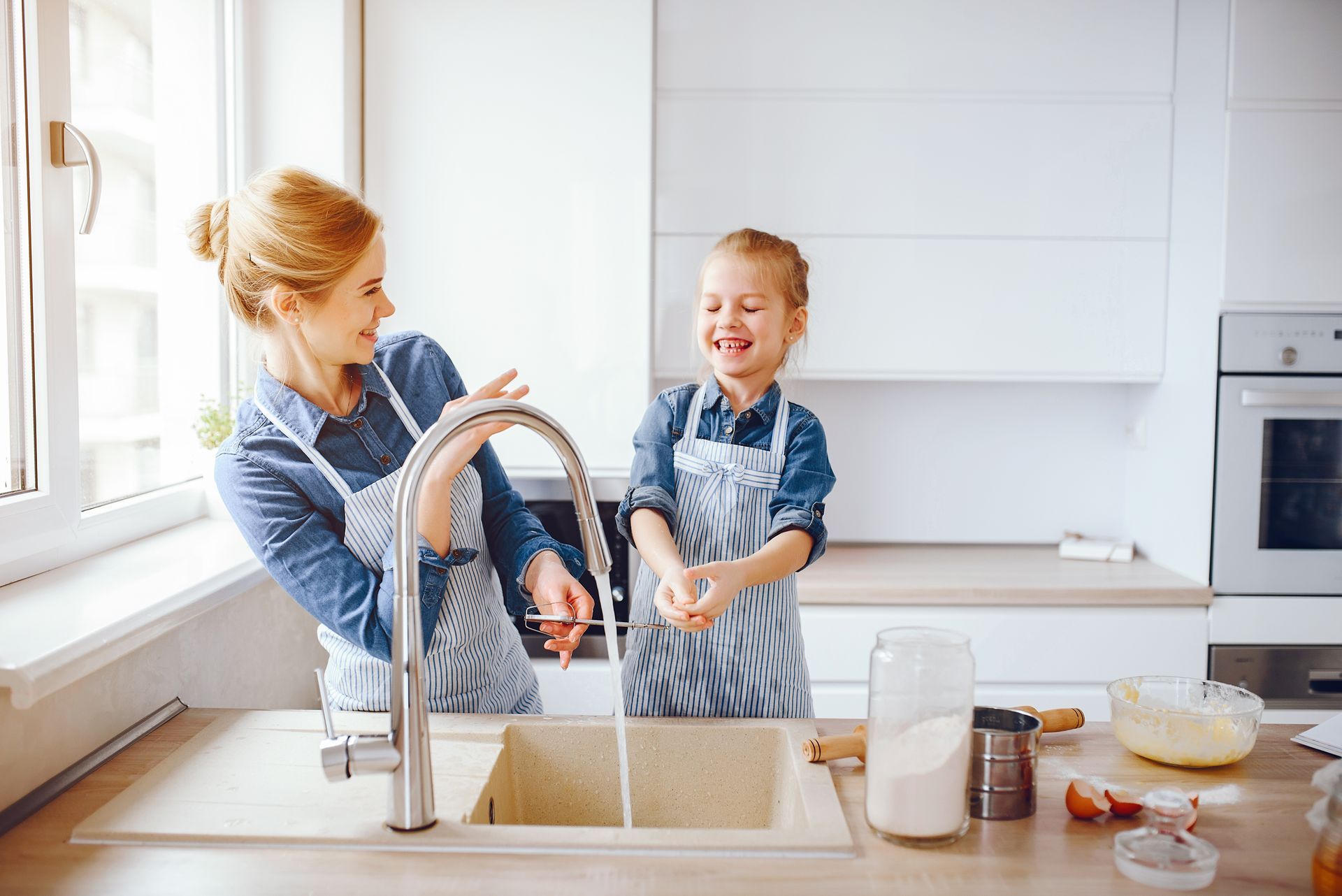 Woman and child washing hands at a kitchen sink, both smiling, wearing aprons.