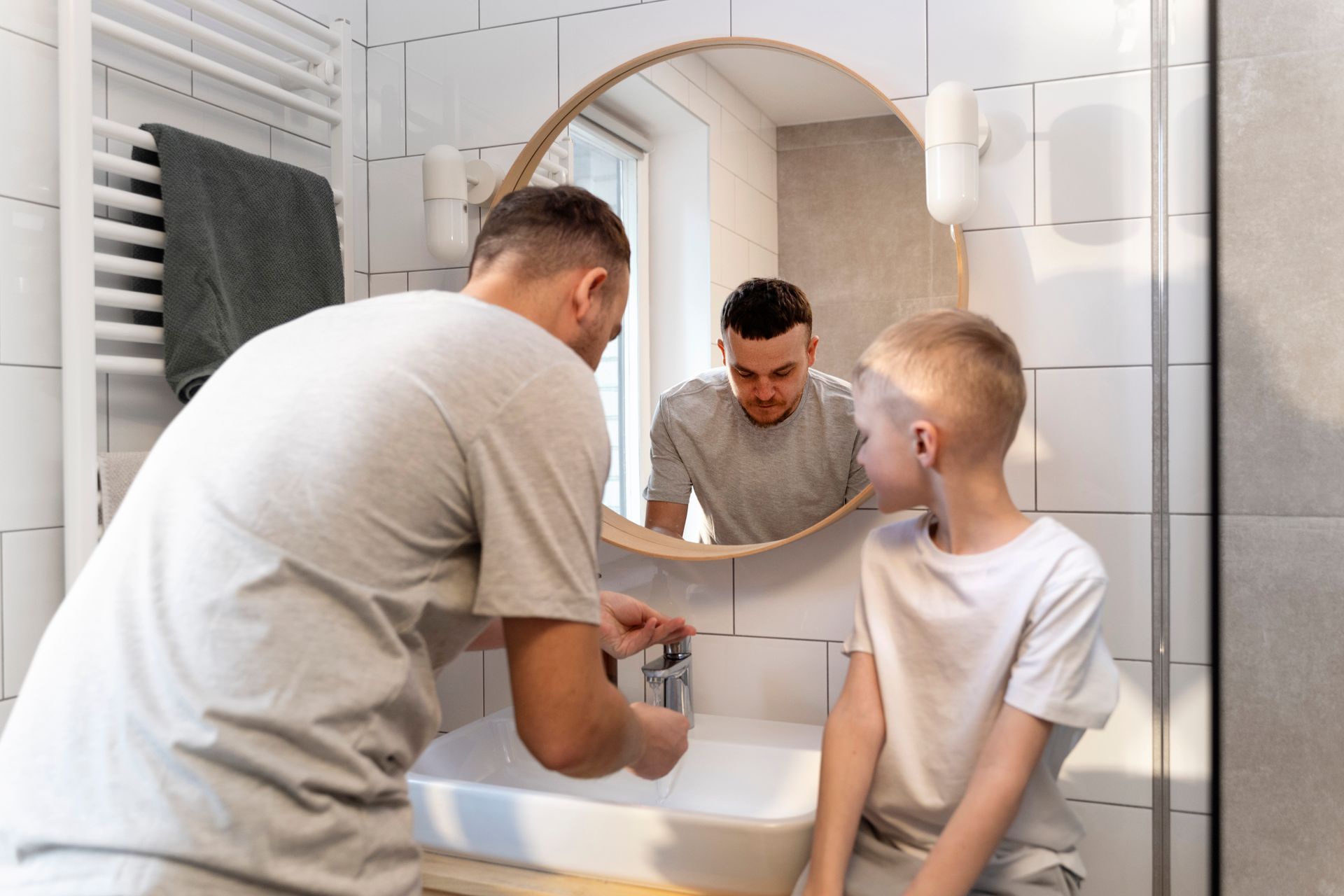 Man and boy washing hands at a bathroom sink, with a circular mirror reflecting them.