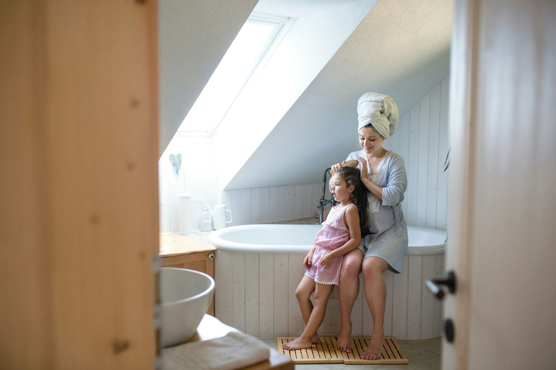 Woman styling a child's hair in a bathroom with a skylight and a white tub.