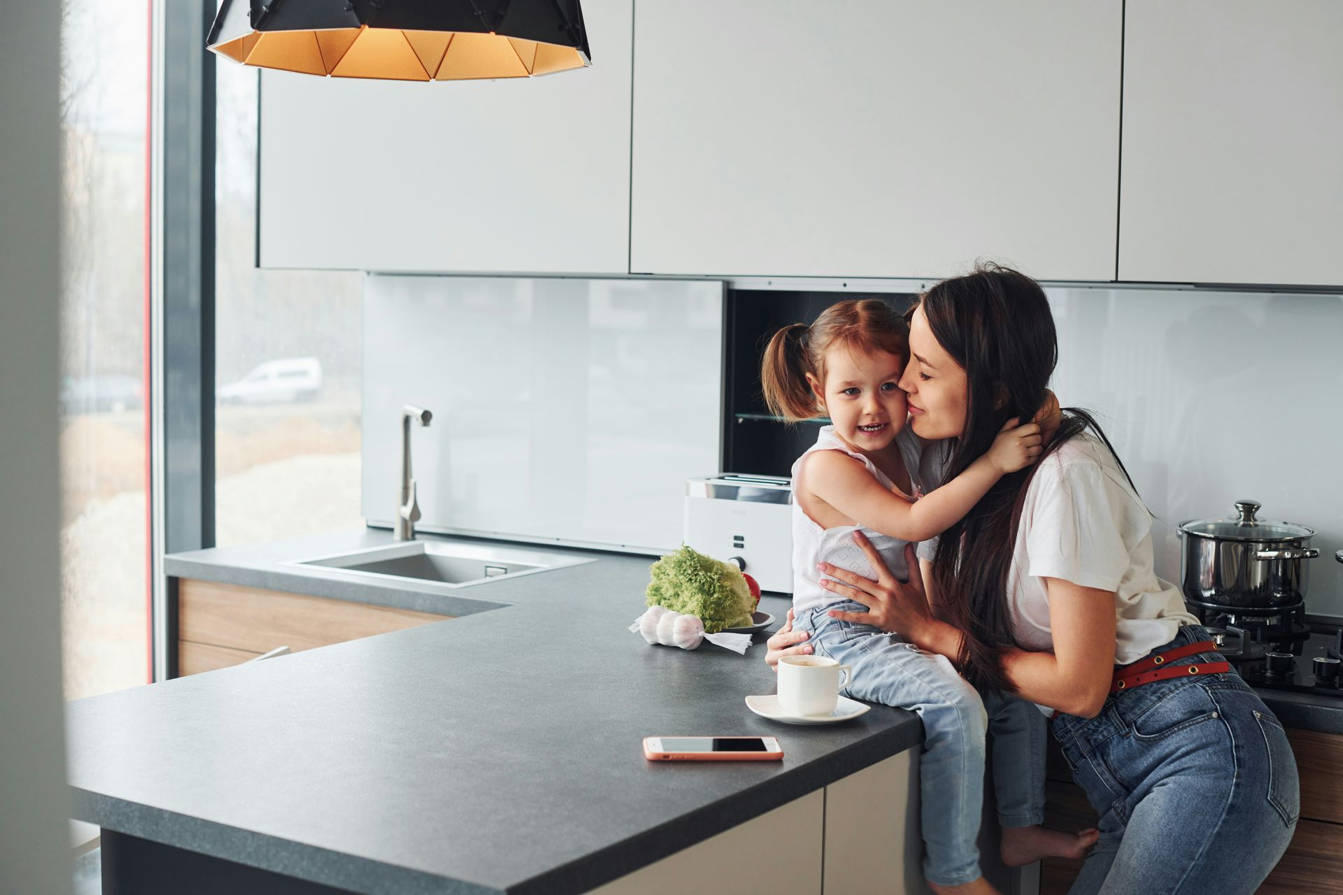 Mother and child embracing in a modern kitchen, with a countertop and overhead lighting.