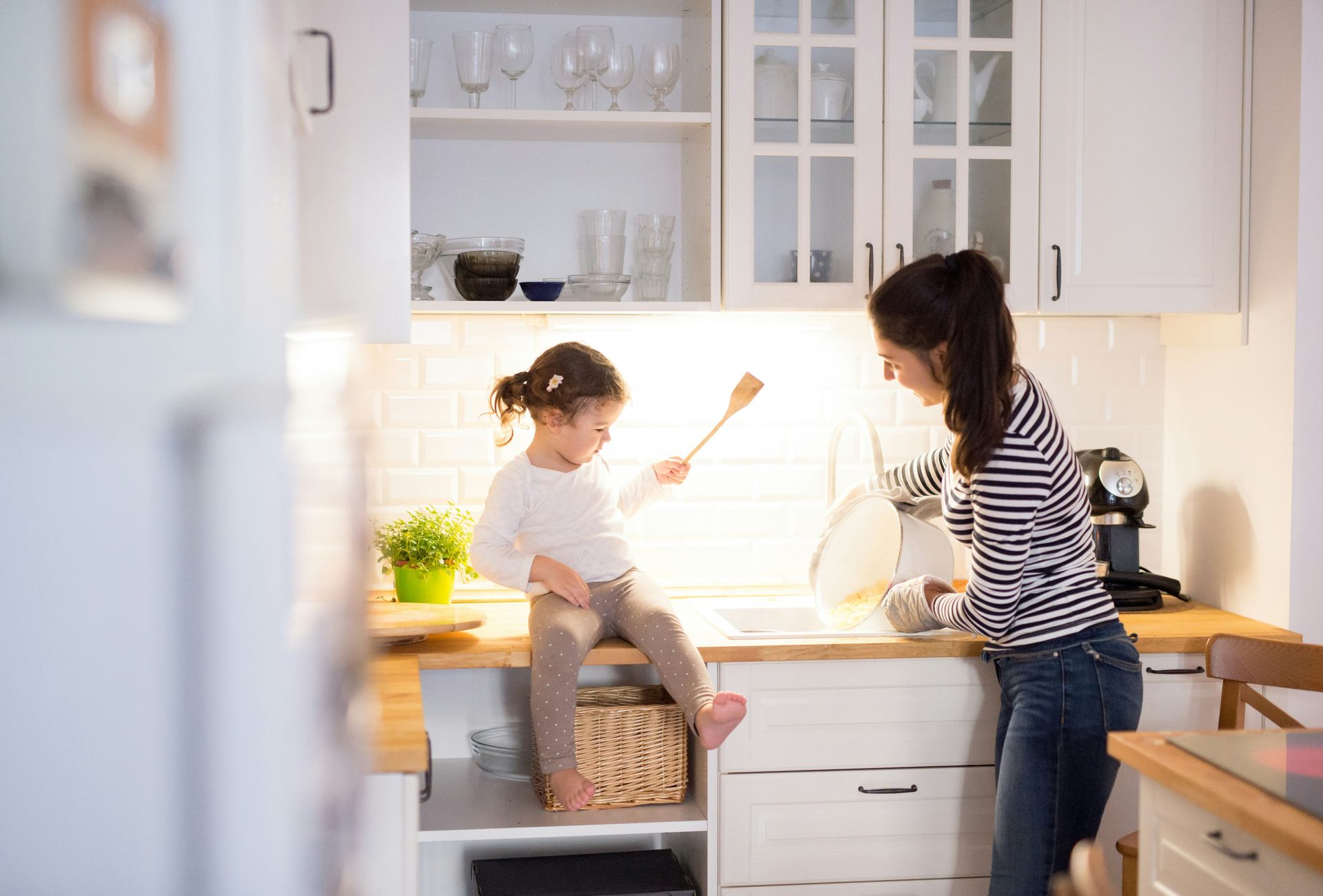 Woman and child in a white kitchen; child on counter, woman washing dishes.