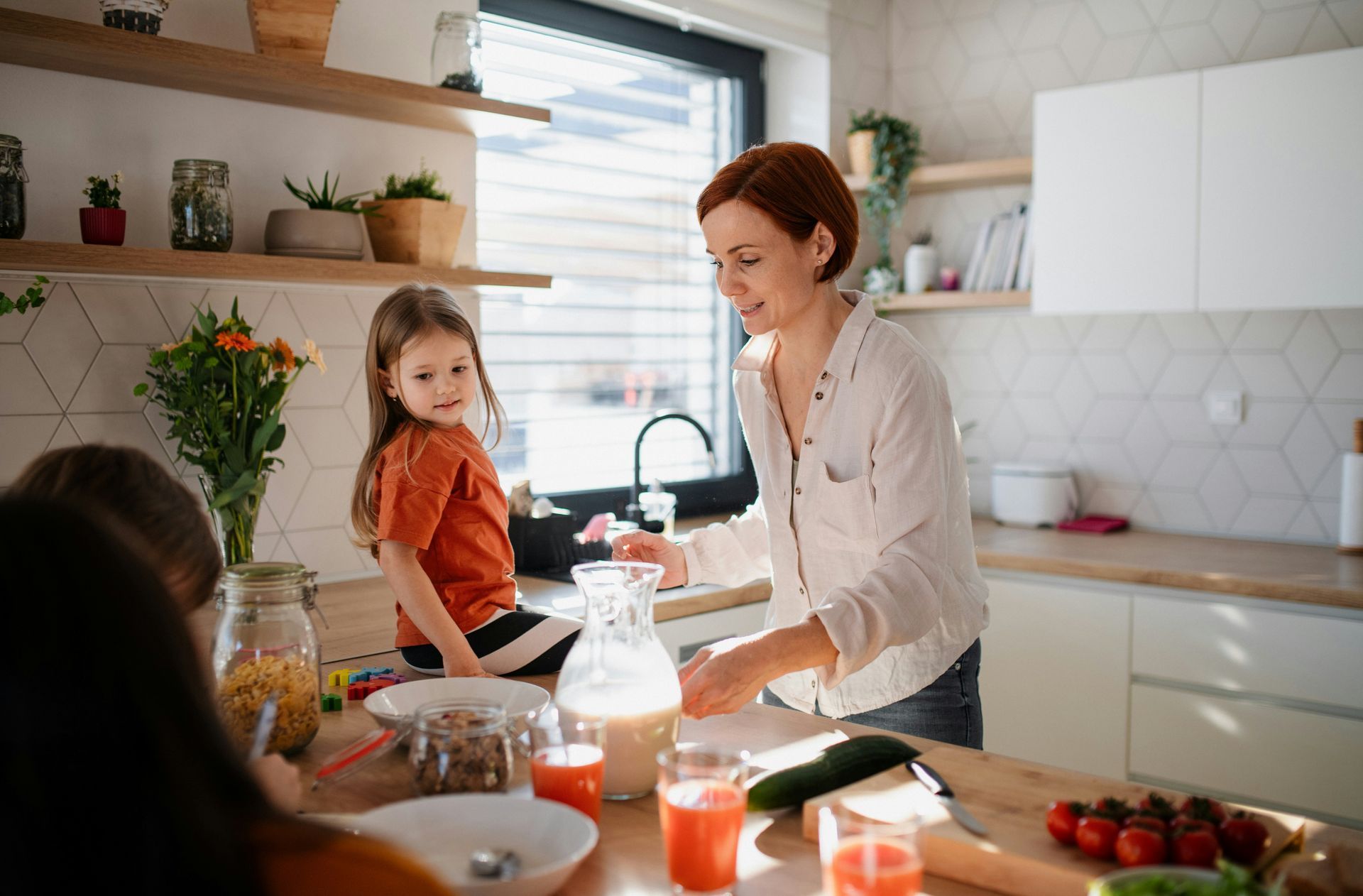 Woman in a kitchen pours liquid as child watches; breakfast items on the table, bright space.