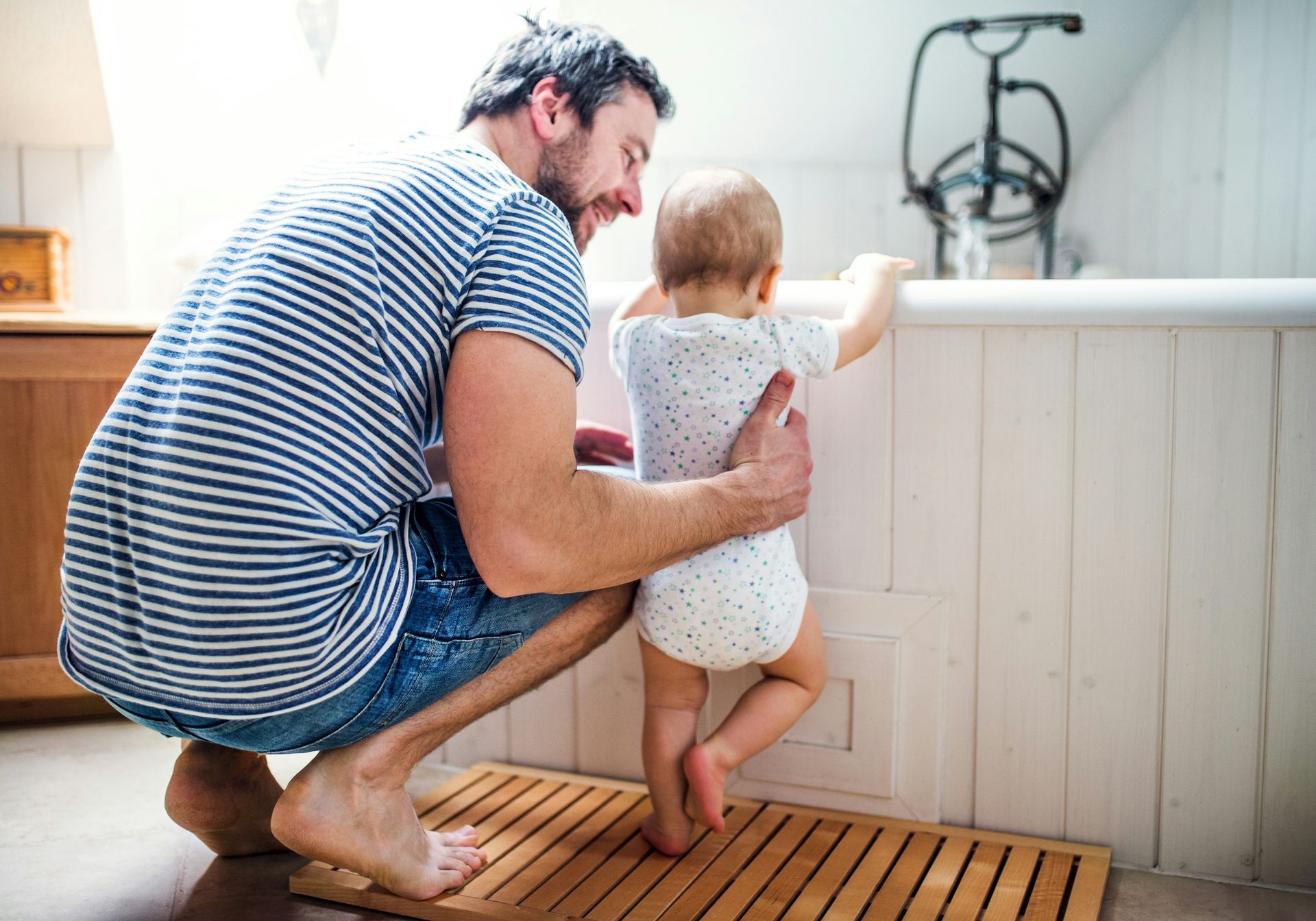 Man helps toddler stand at a bathtub. They are in a bathroom.