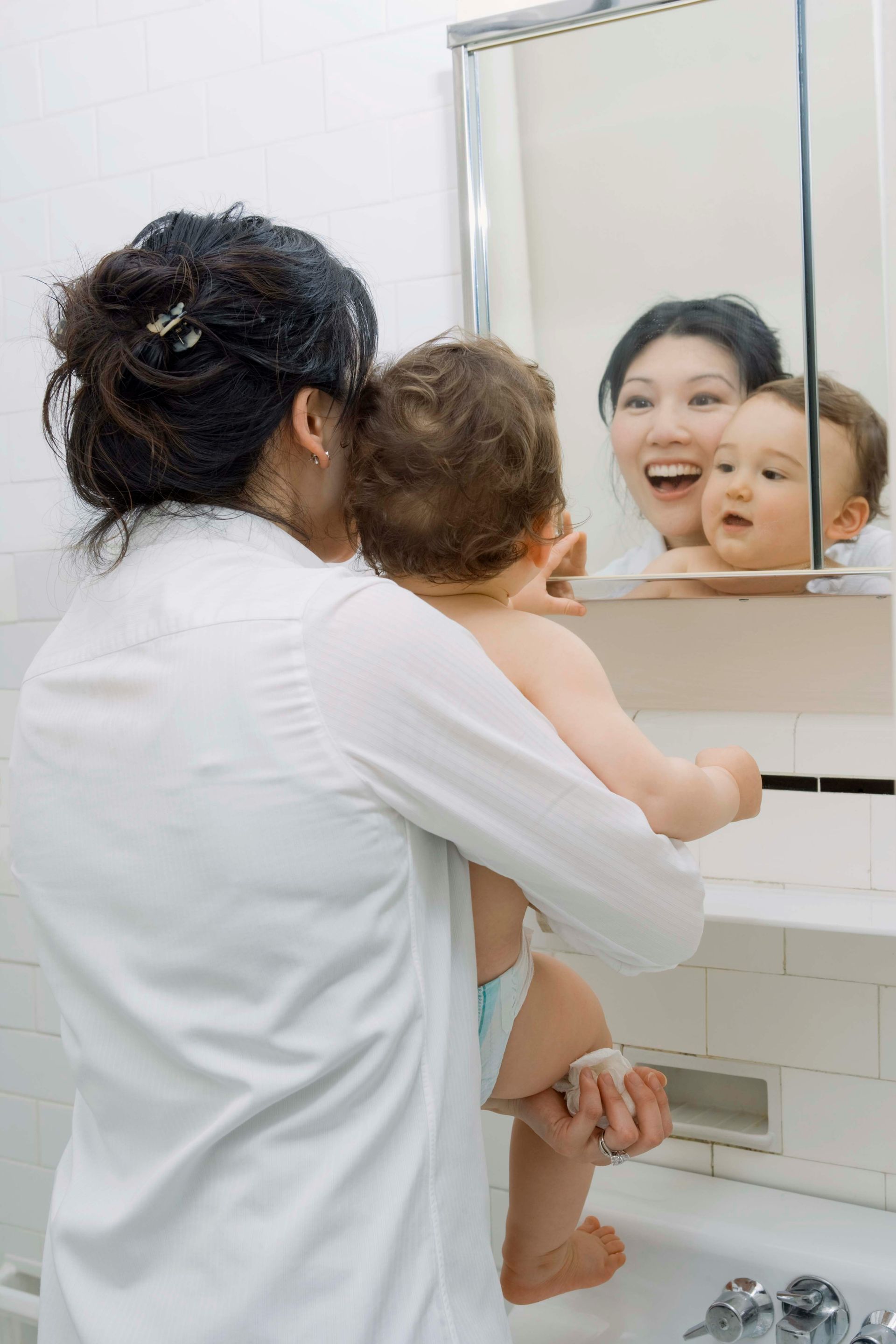 Woman holding baby in front of a mirror, baby looking at reflection. Woman smiling. Bathroom setting.