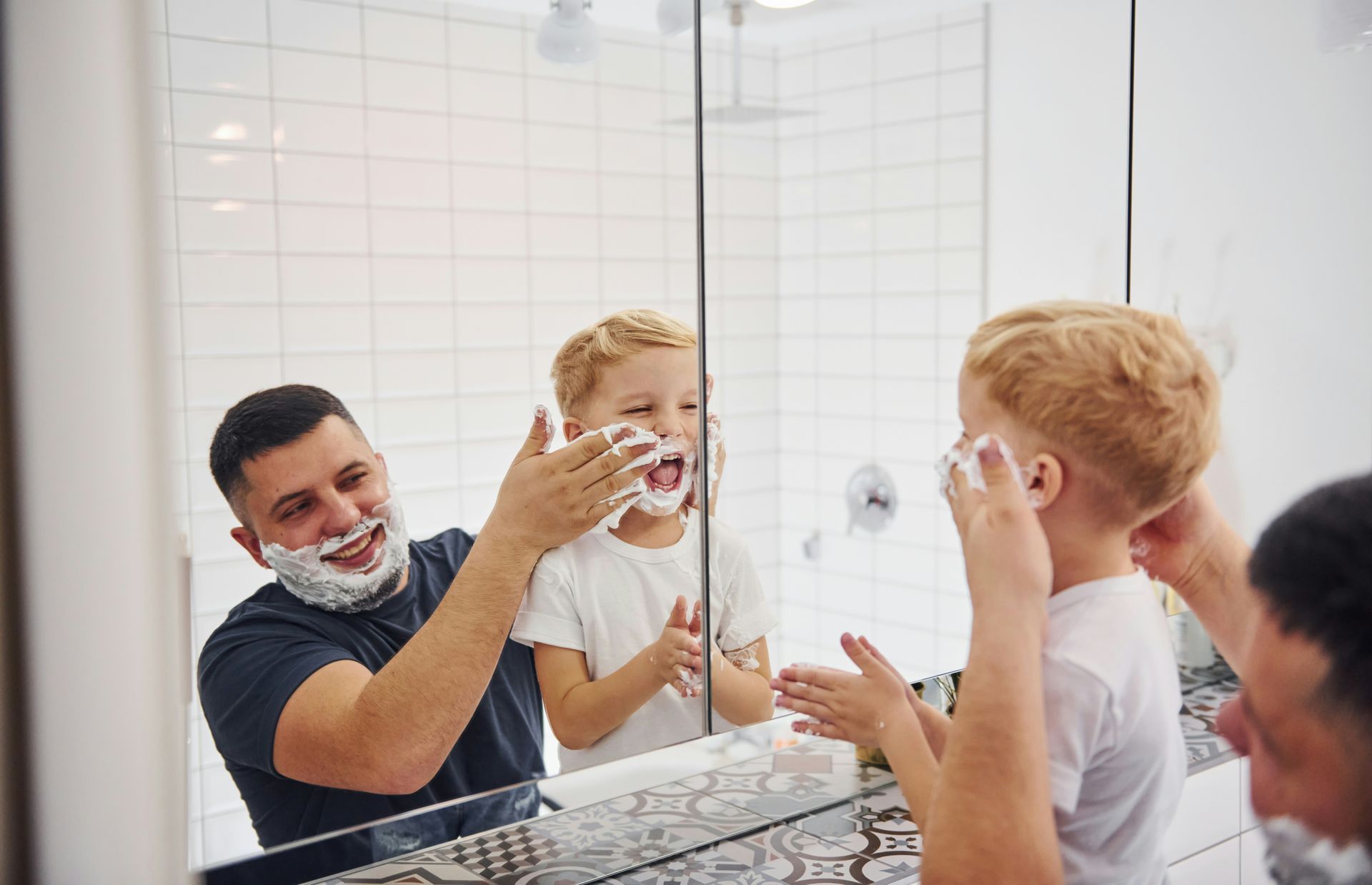 Man and child applying shaving cream in front of a mirror in a bathroom; both are smiling.