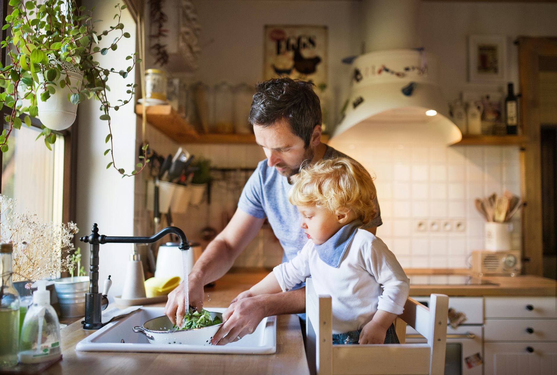 Man and child in kitchen, man washing vegetables, child in a learning tower watching.