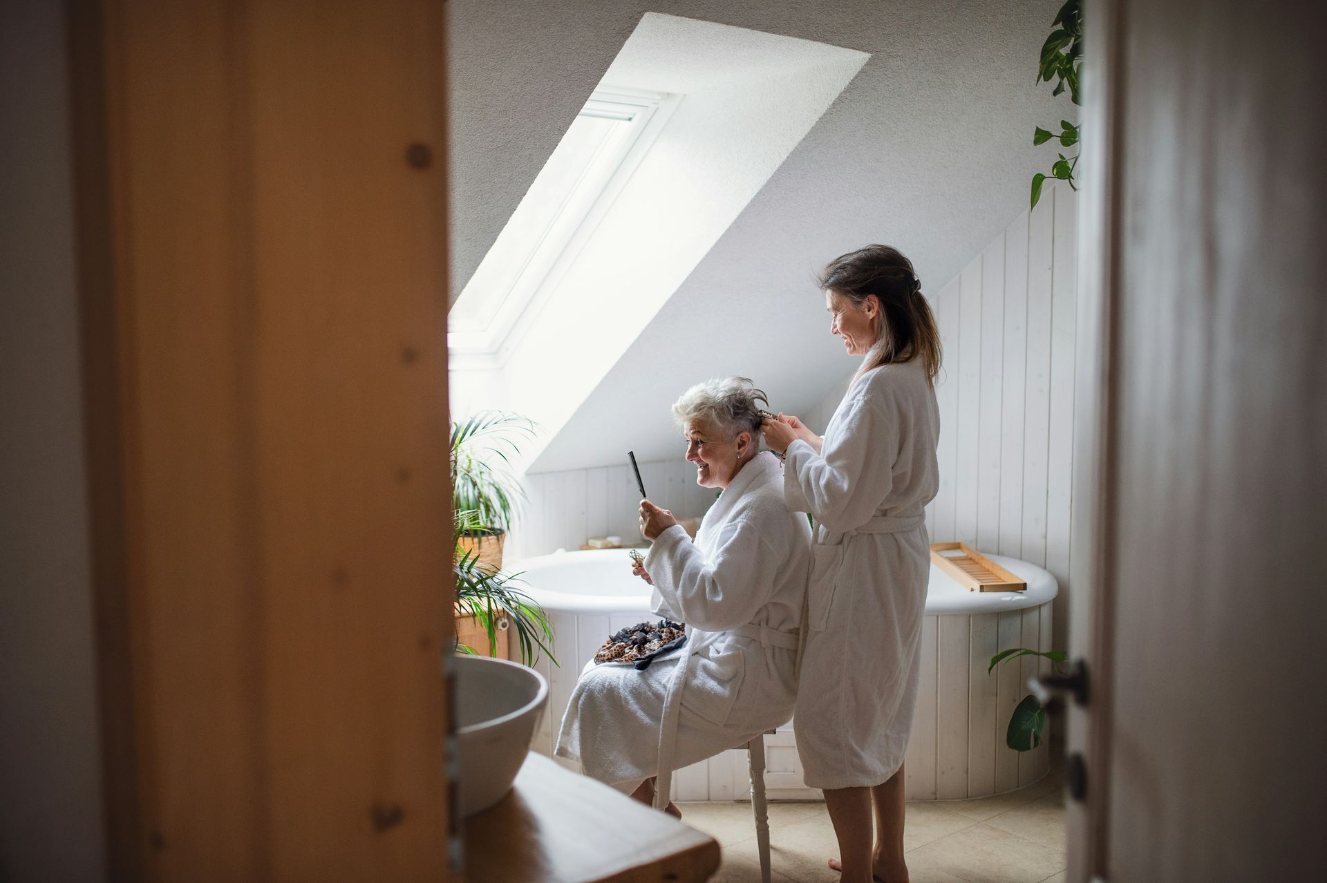 Woman helping another woman with her hair in a bathroom with a skylight and a bathtub.