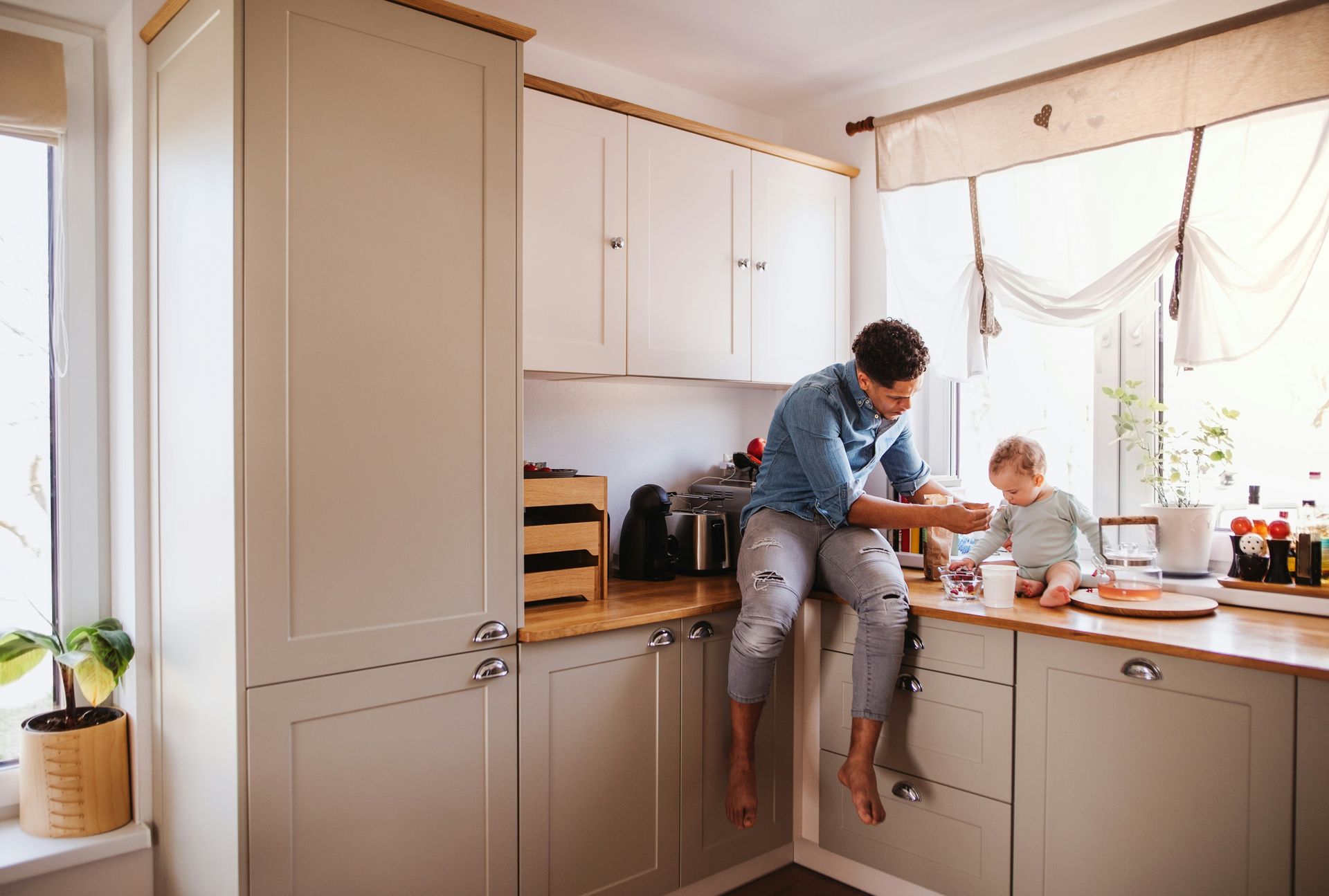 Man and toddler sit on kitchen counter; man reaches, child watches.
