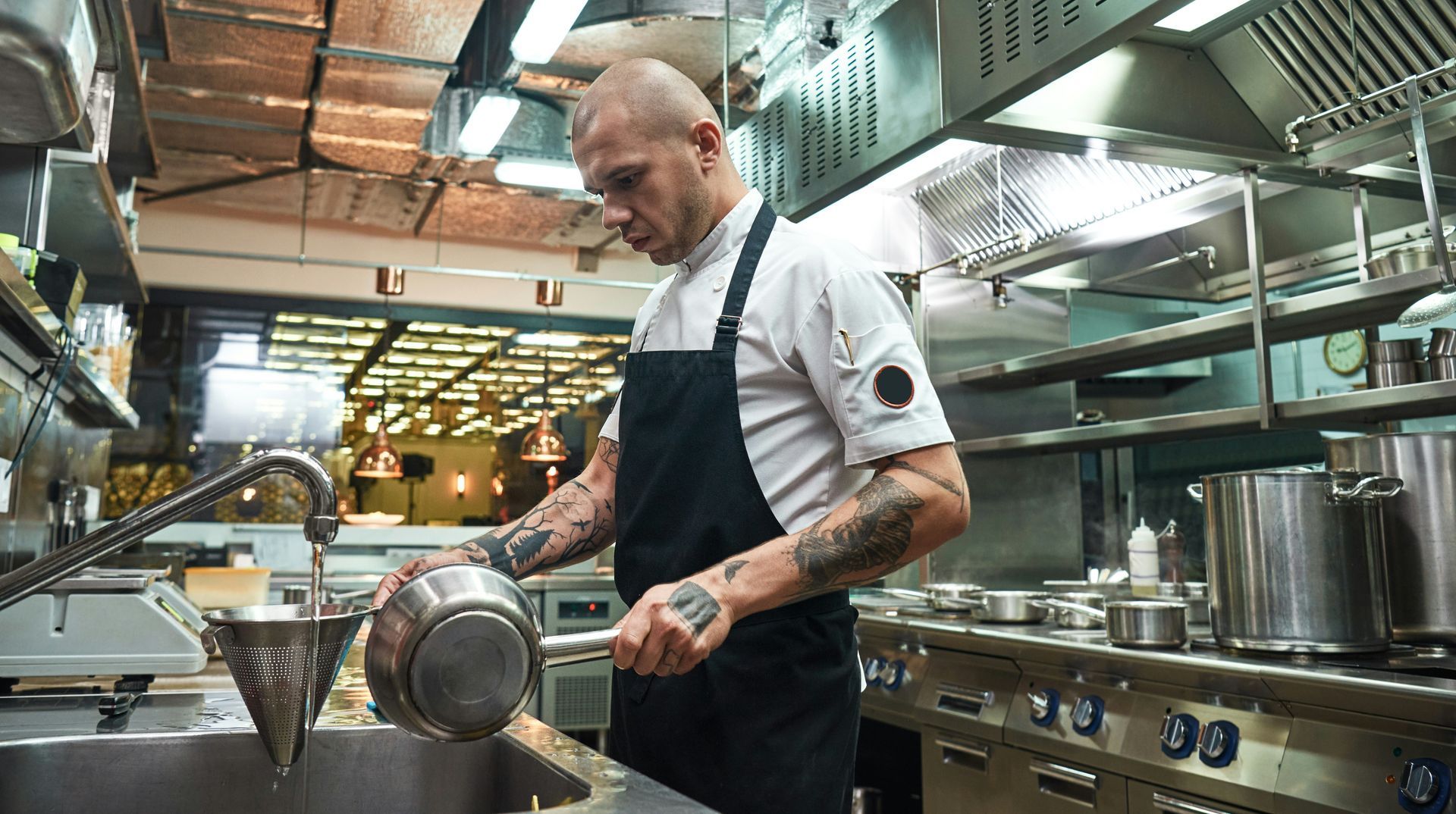 Chef pouring from a pot into a strainer over a sink in a commercial kitchen.