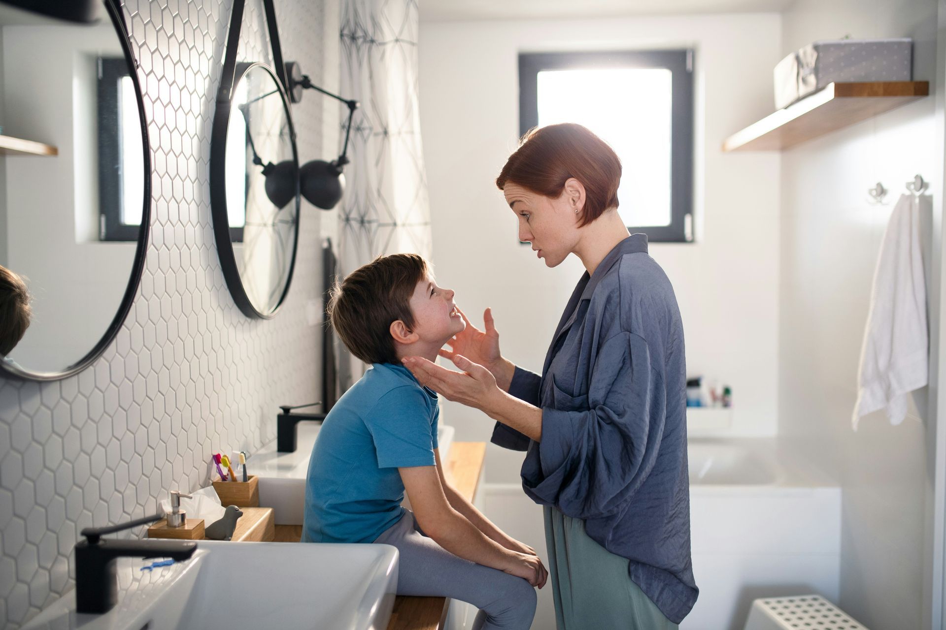 Woman checks boy's face in bathroom. White tiled wall, two mirrors, sunlight.