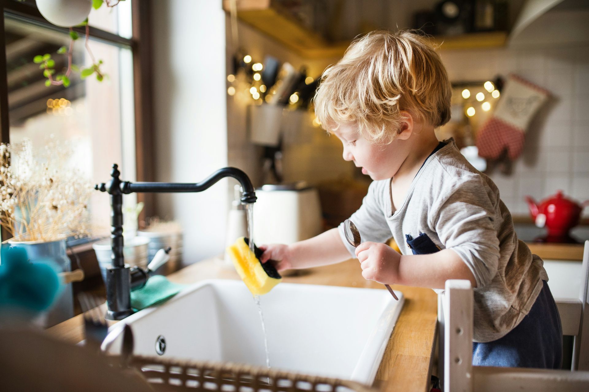 Child washing dishes at kitchen sink. Water running, sponge in hand, wooden counter.