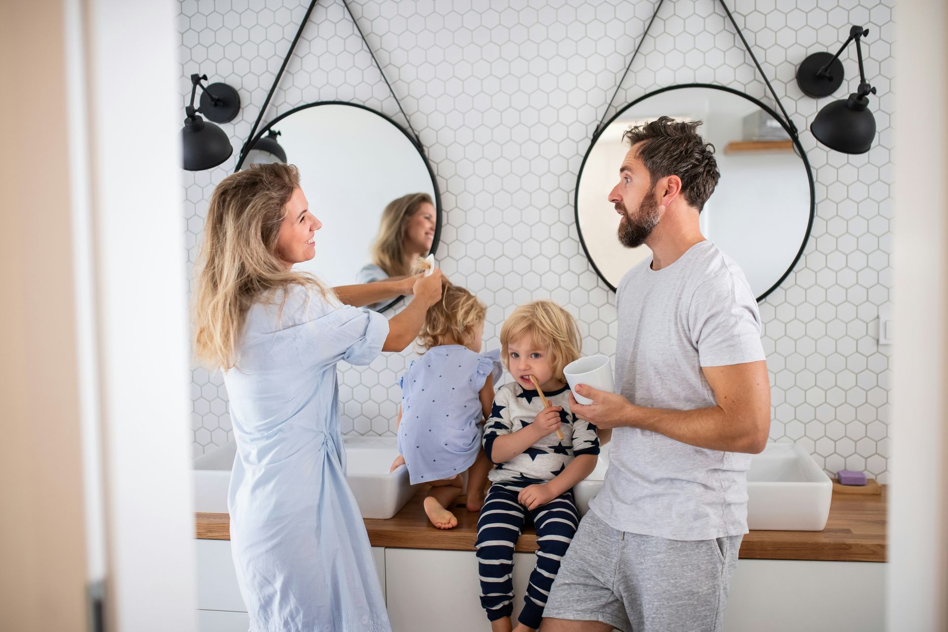 Family brushing teeth in a modern bathroom. Mother combs son's hair, father brushes, siblings watch.