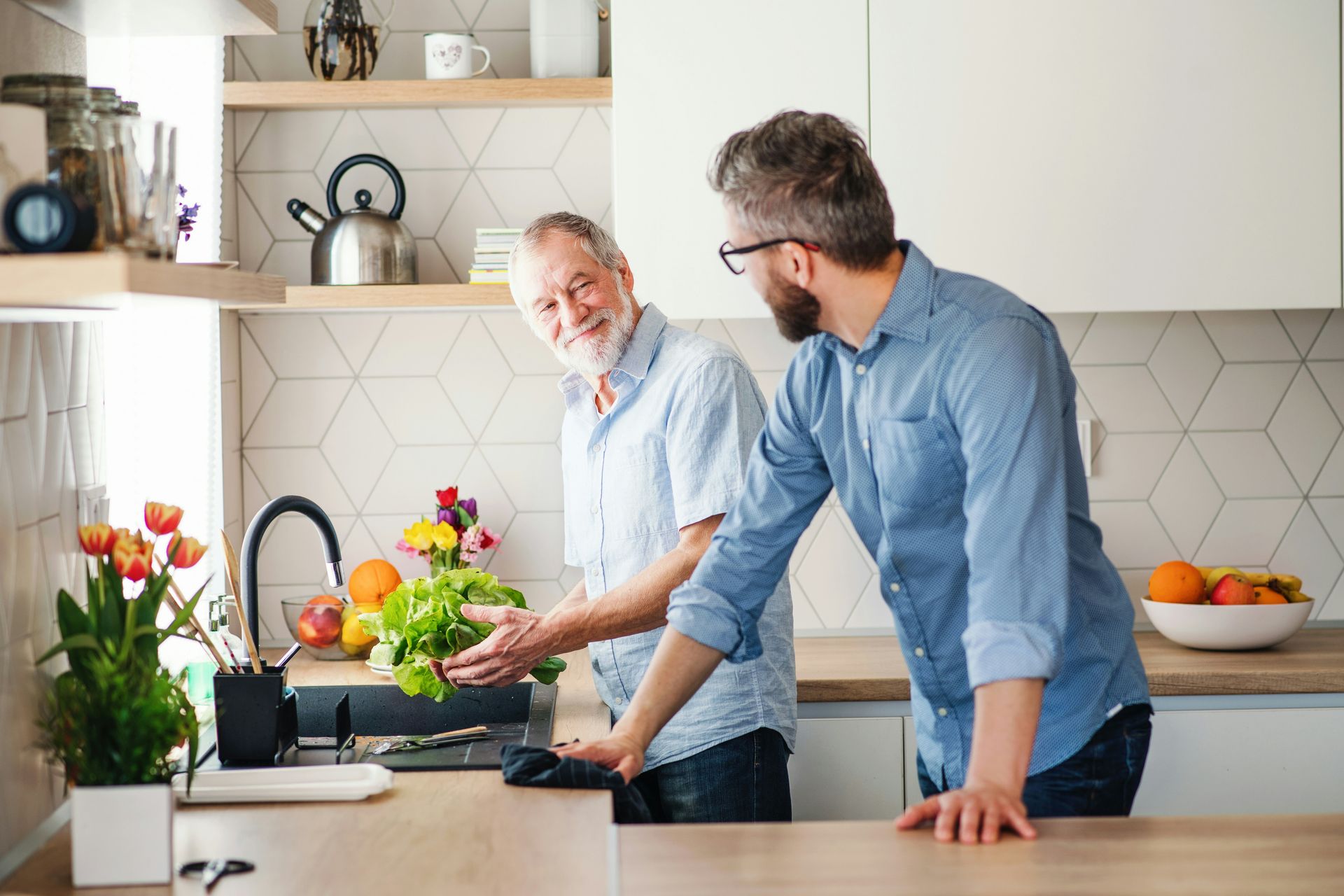 Two men in kitchen, one washing lettuce, smiling. Modern, light-filled kitchen.