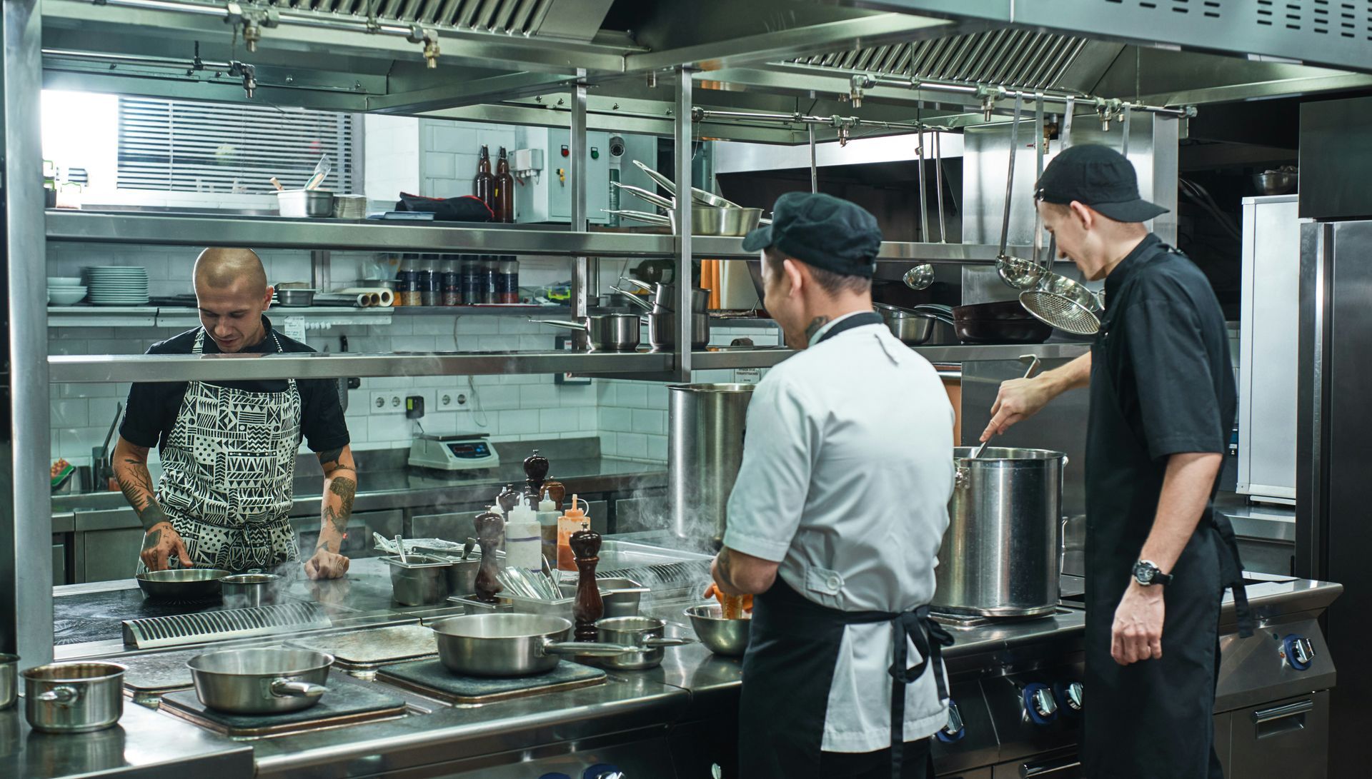 Three chefs working in a commercial kitchen; one stirring a pot, one preparing food, and one behind a counter.