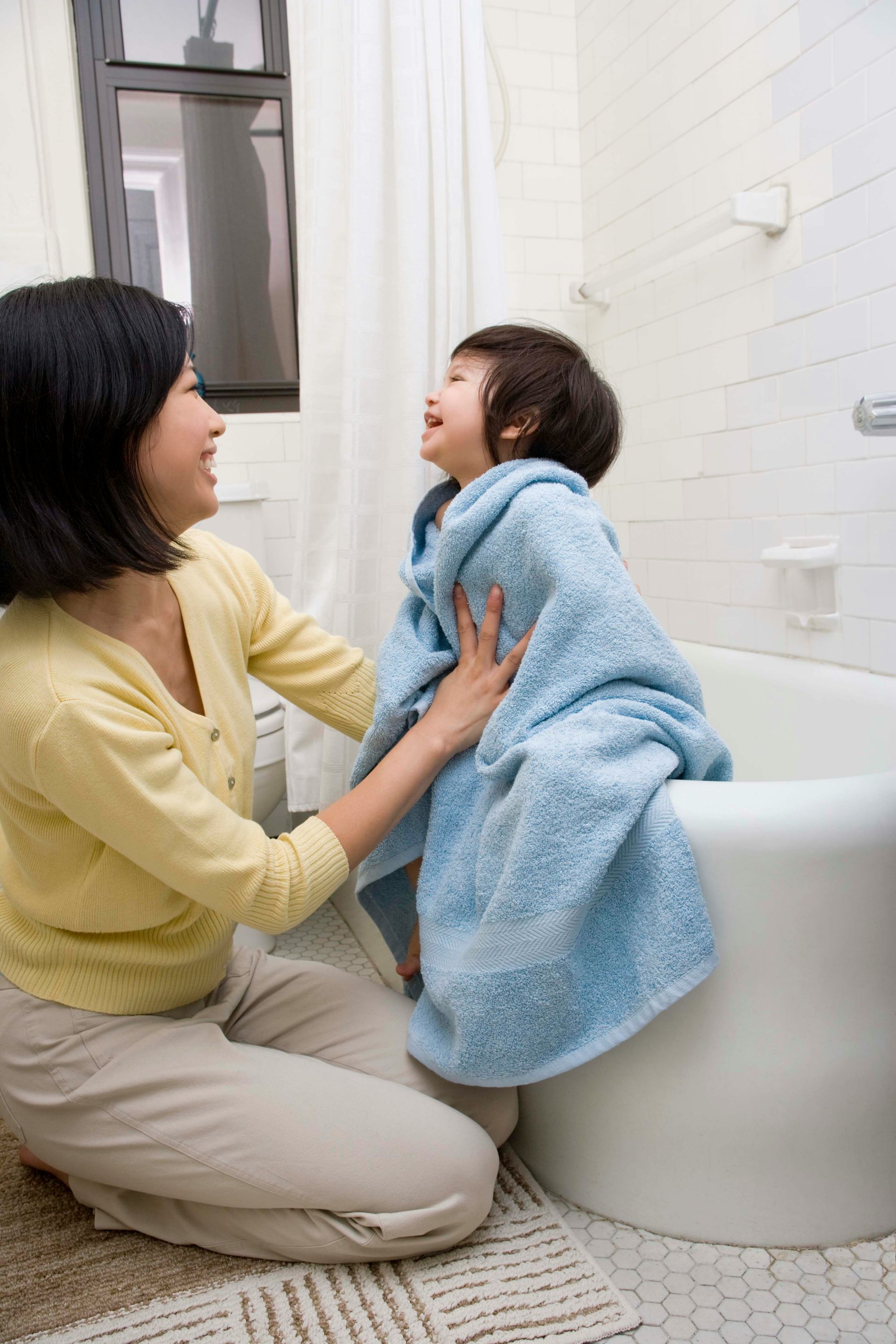 Woman wrapping toddler in a blue towel after a bath, both smiling in a bright bathroom.