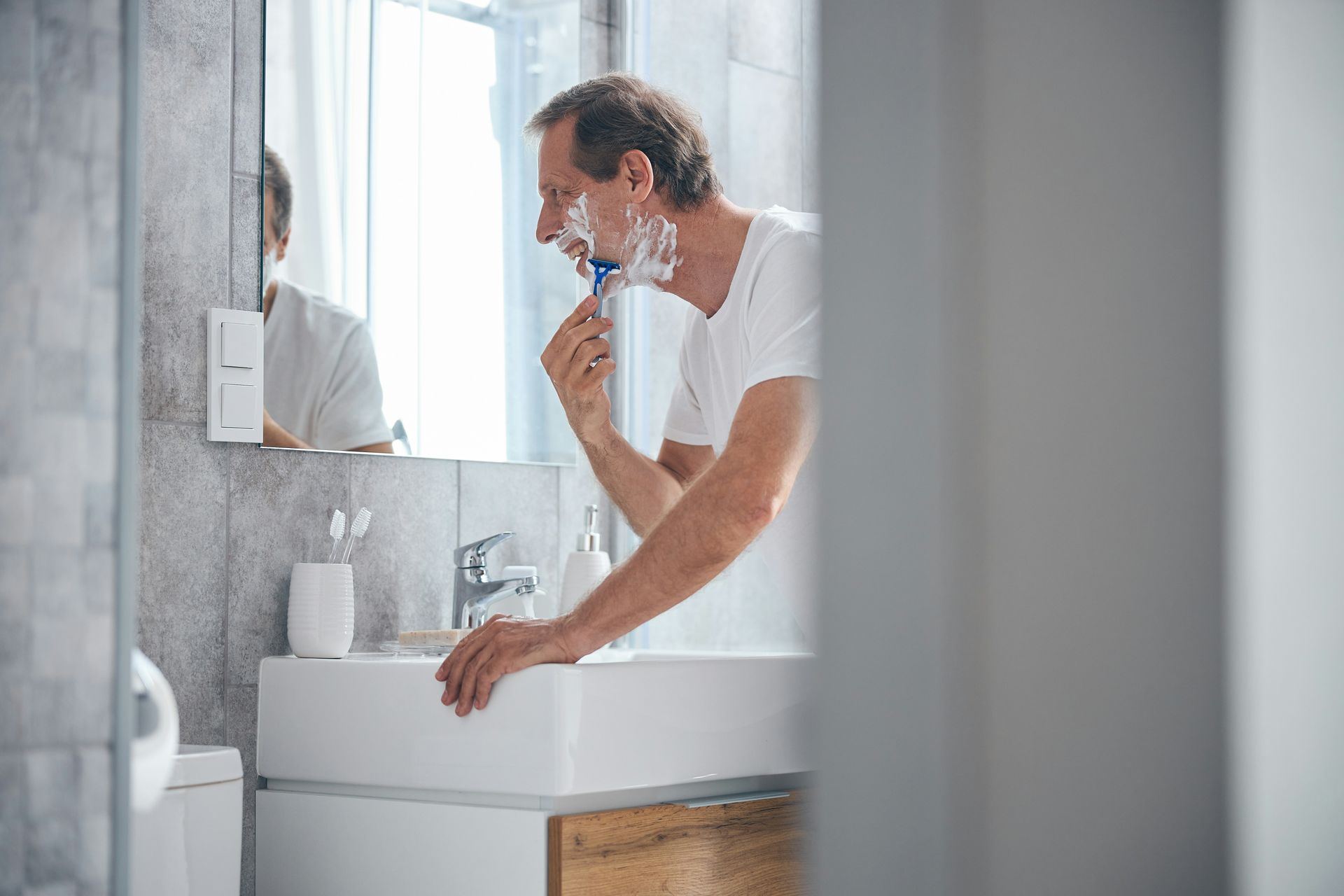 Man shaving in bathroom, using razor. White sink, mirror reflection.