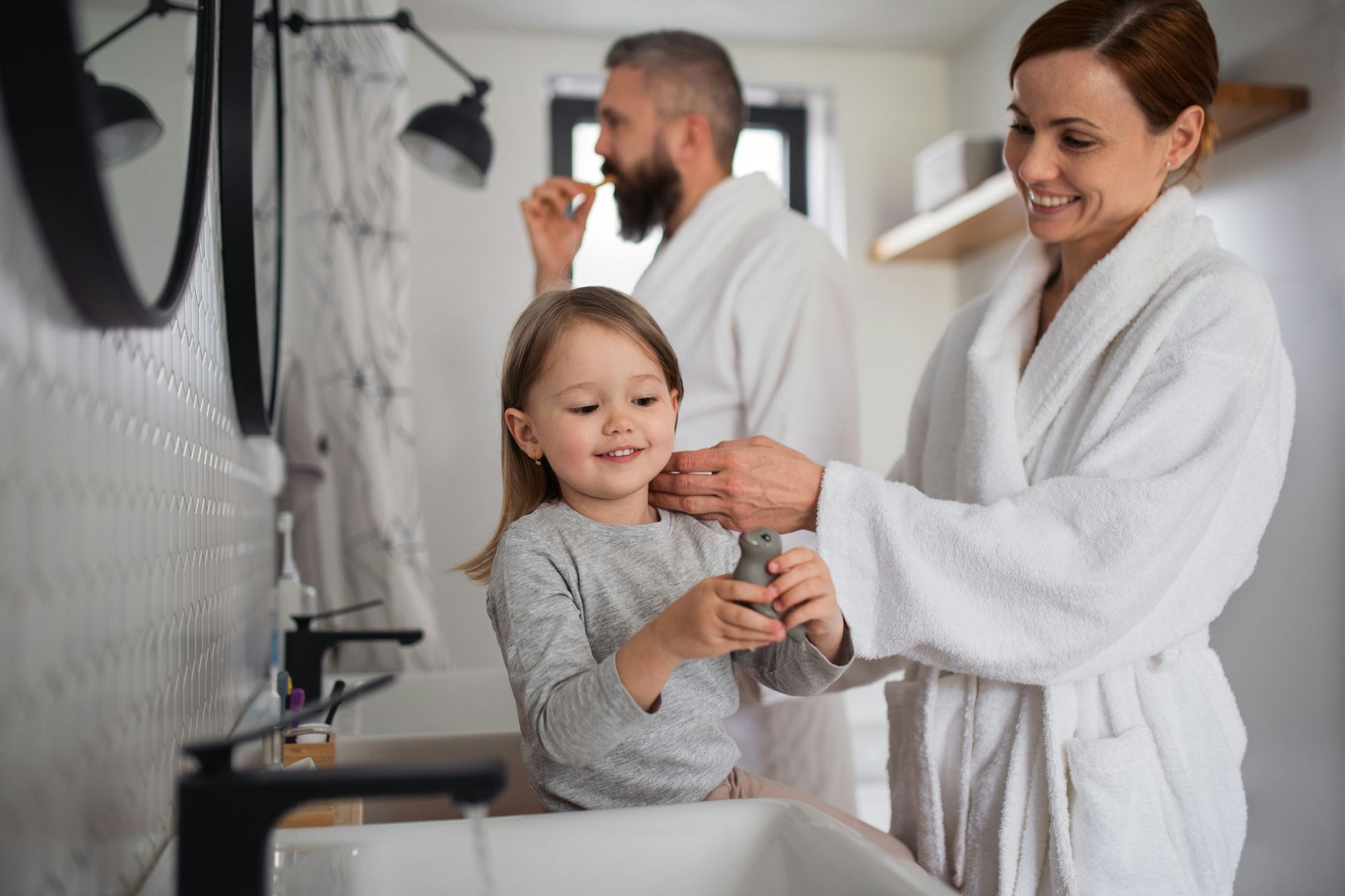 Family in bathroom brushing teeth: a smiling woman helps a child while a man brushes his teeth in background.